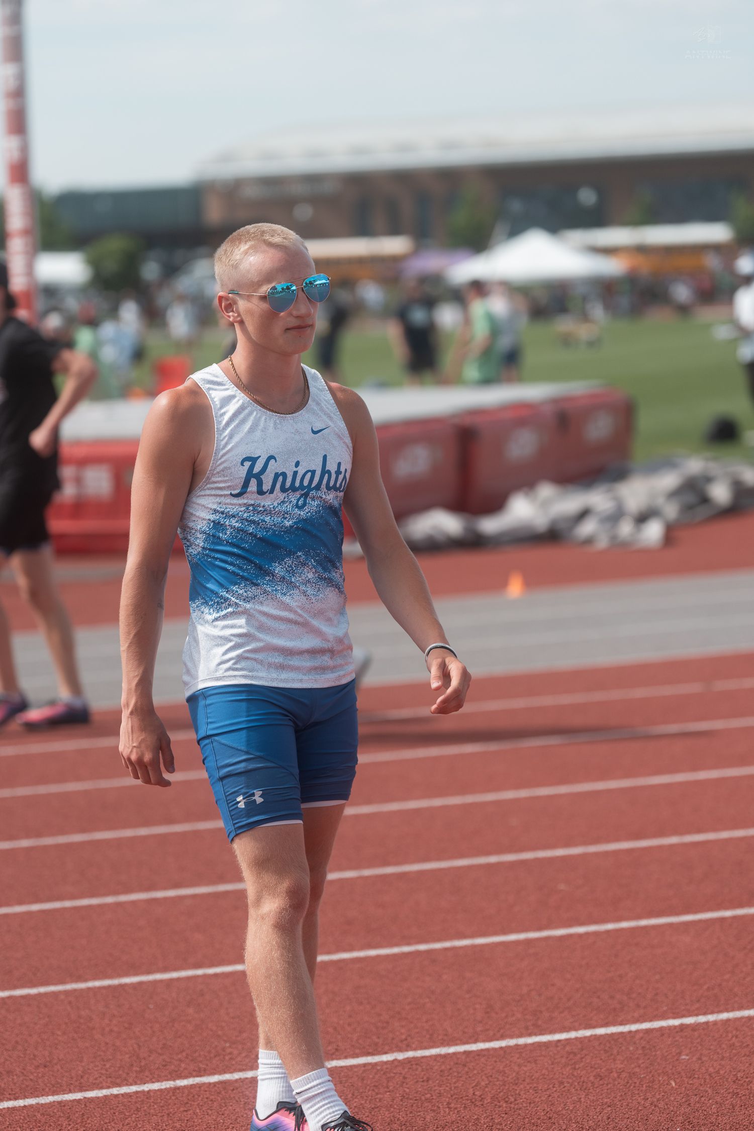 Male Knights sprinter wearing sunglasses walks on red track at sunny outdoor track and field event.
