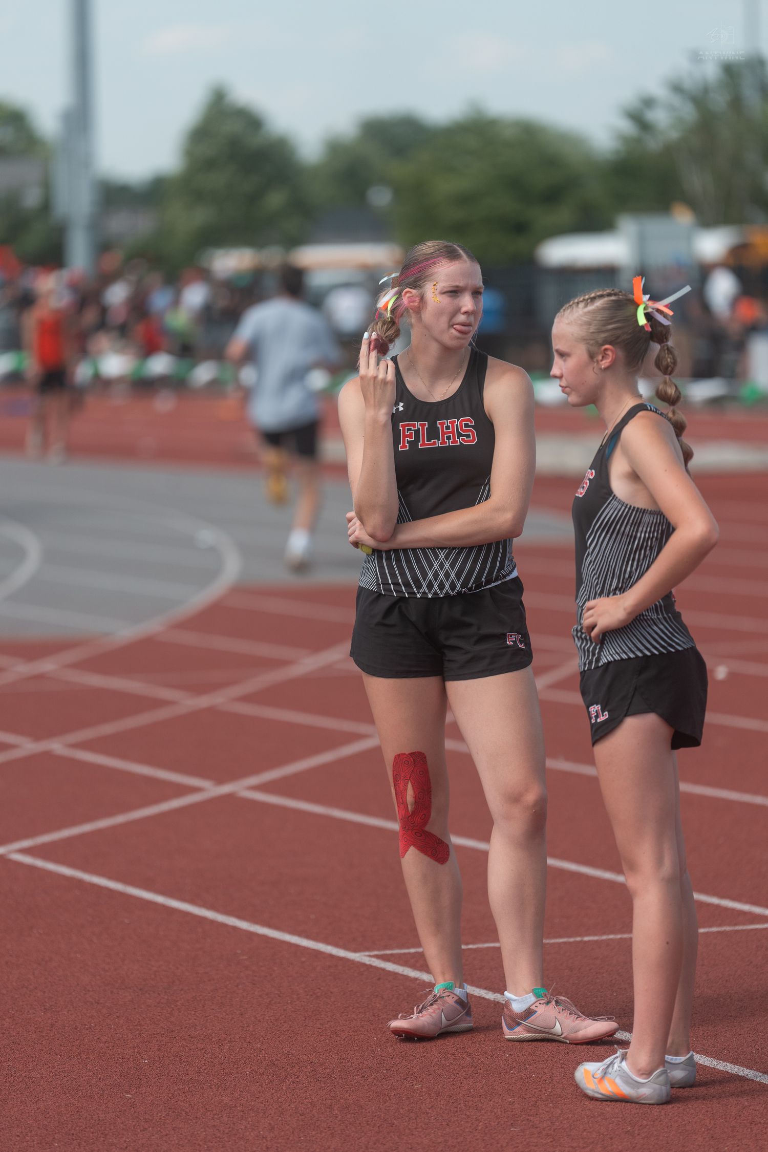 Two female track athletes in FLBS uniforms converse on a red track during a meet.