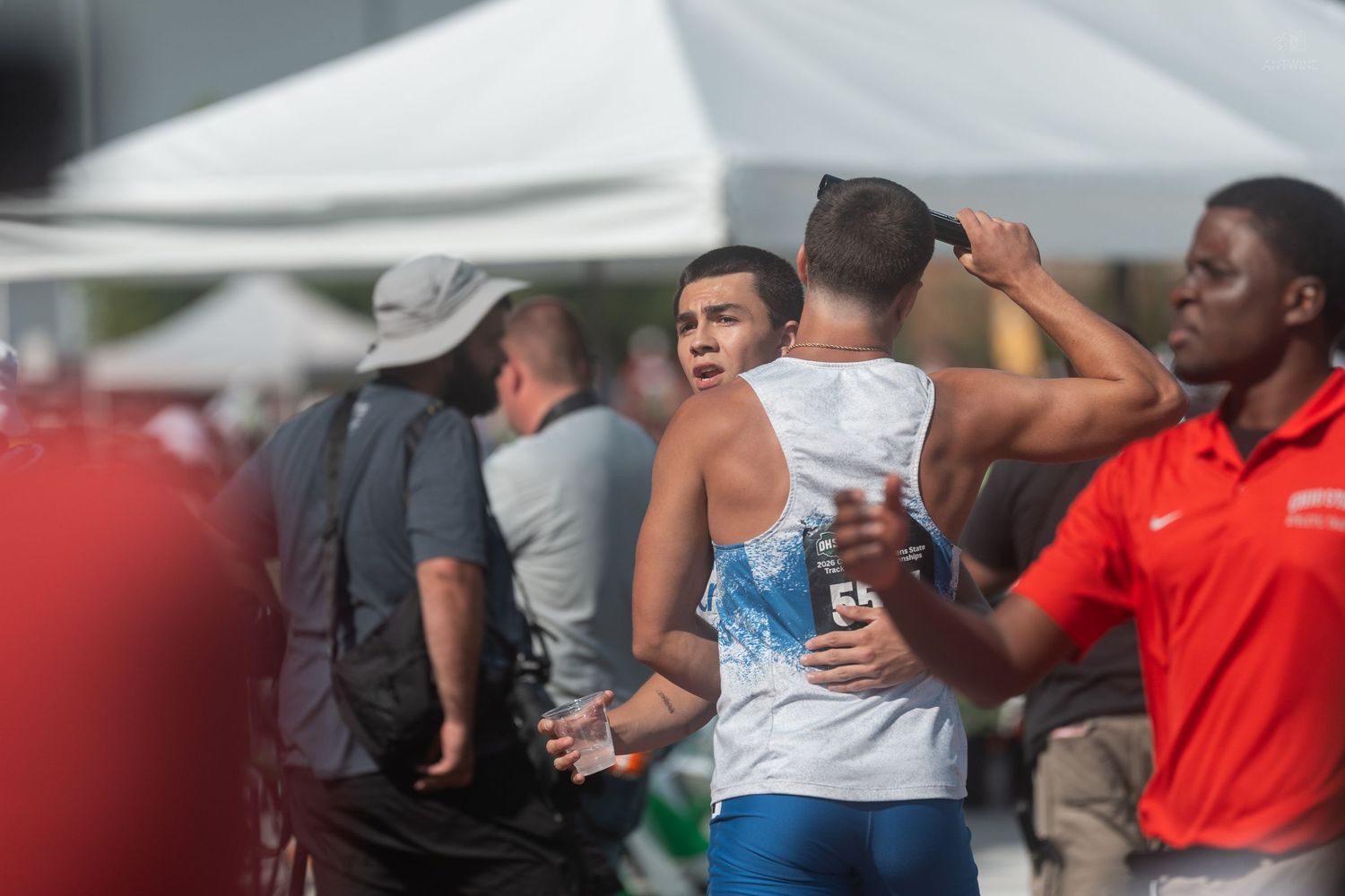 Young athletes share an emotional hug at an outdoor track and field meet with spectators in the background.
