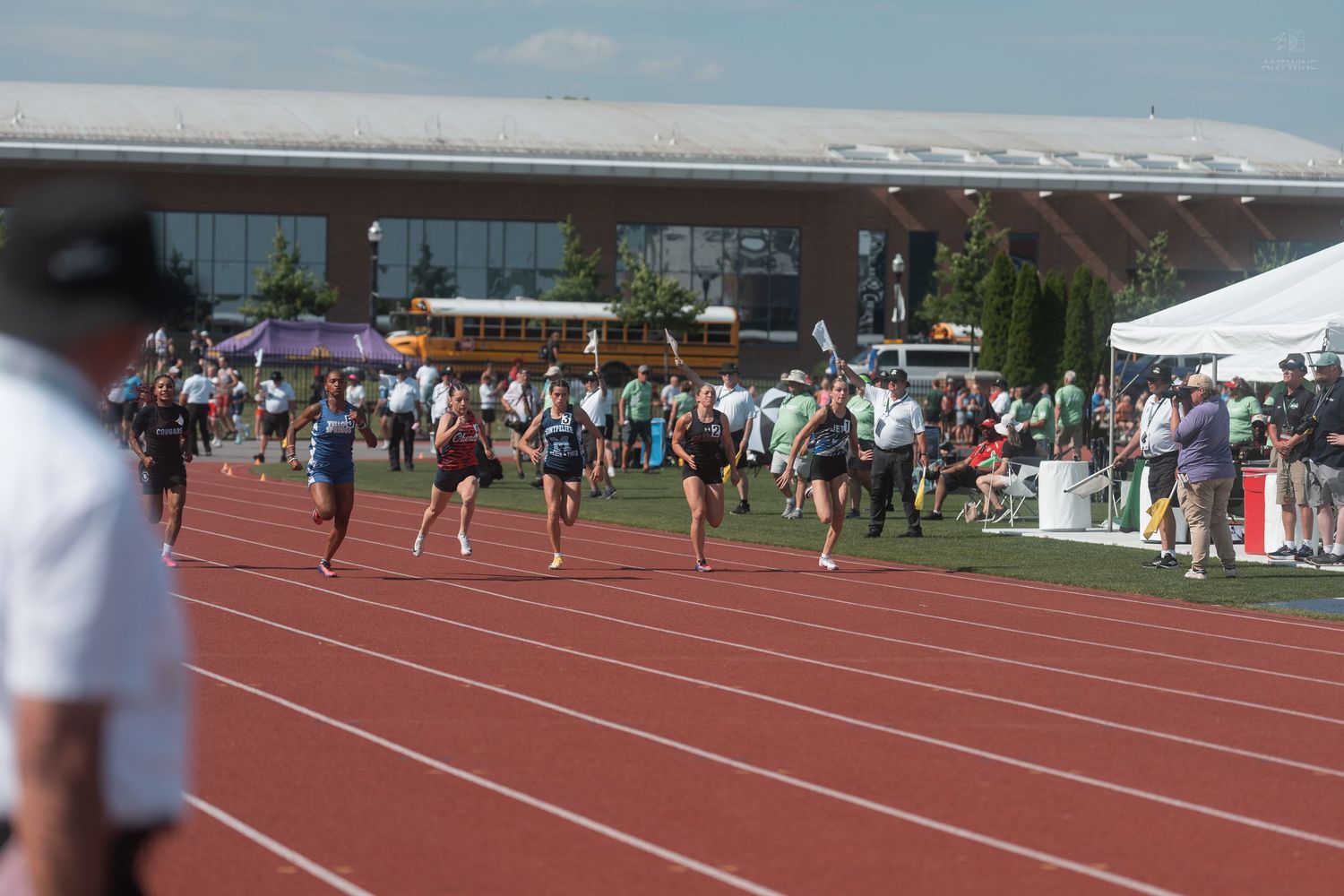 Female track athletes run on a red outdoor track with a school bus and building visible in background.
