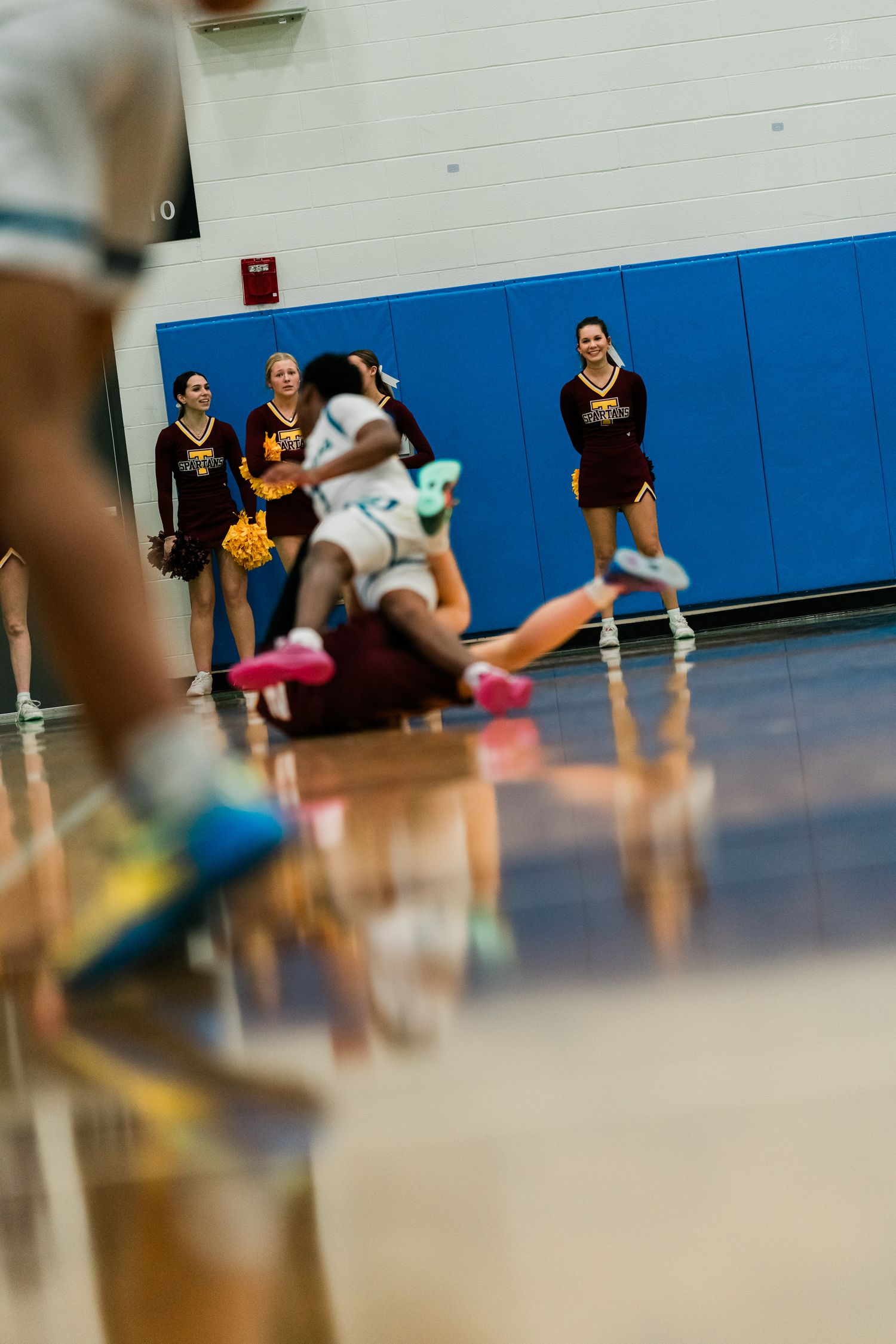 Cheerleaders in maroon uniforms watch as a basketball player falls to the court during a high school game.