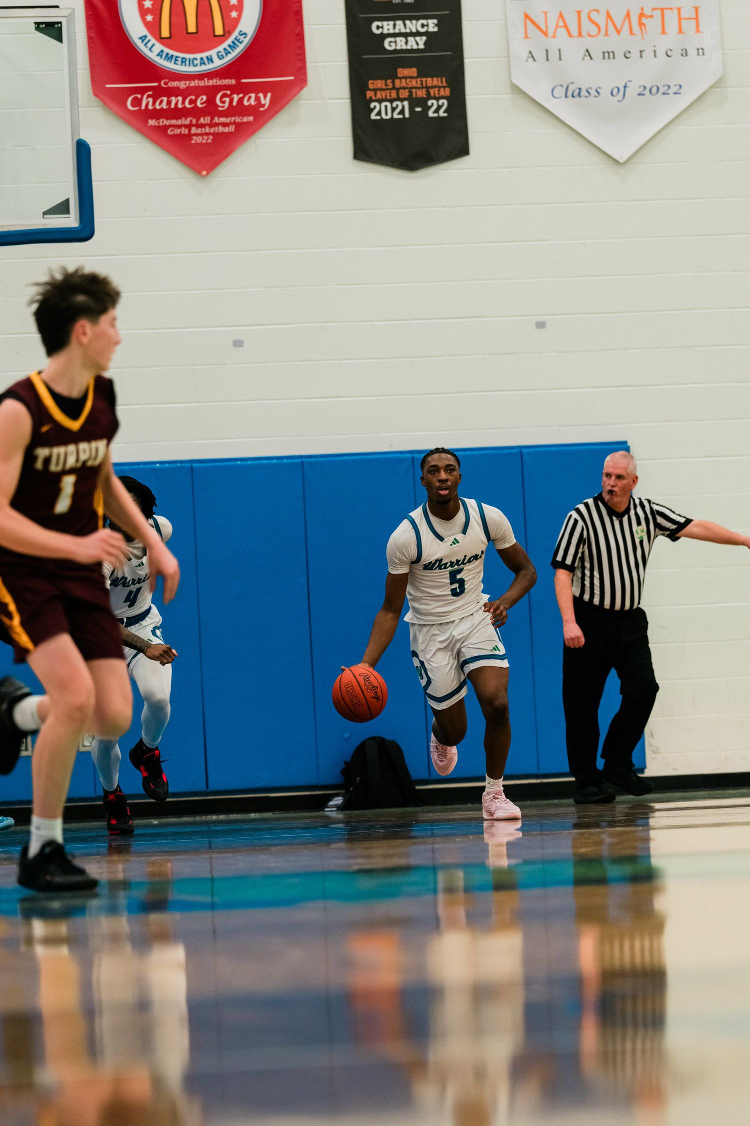 A player in white dribbles the basketball upcourt during a high school game as a referee watches nearby.