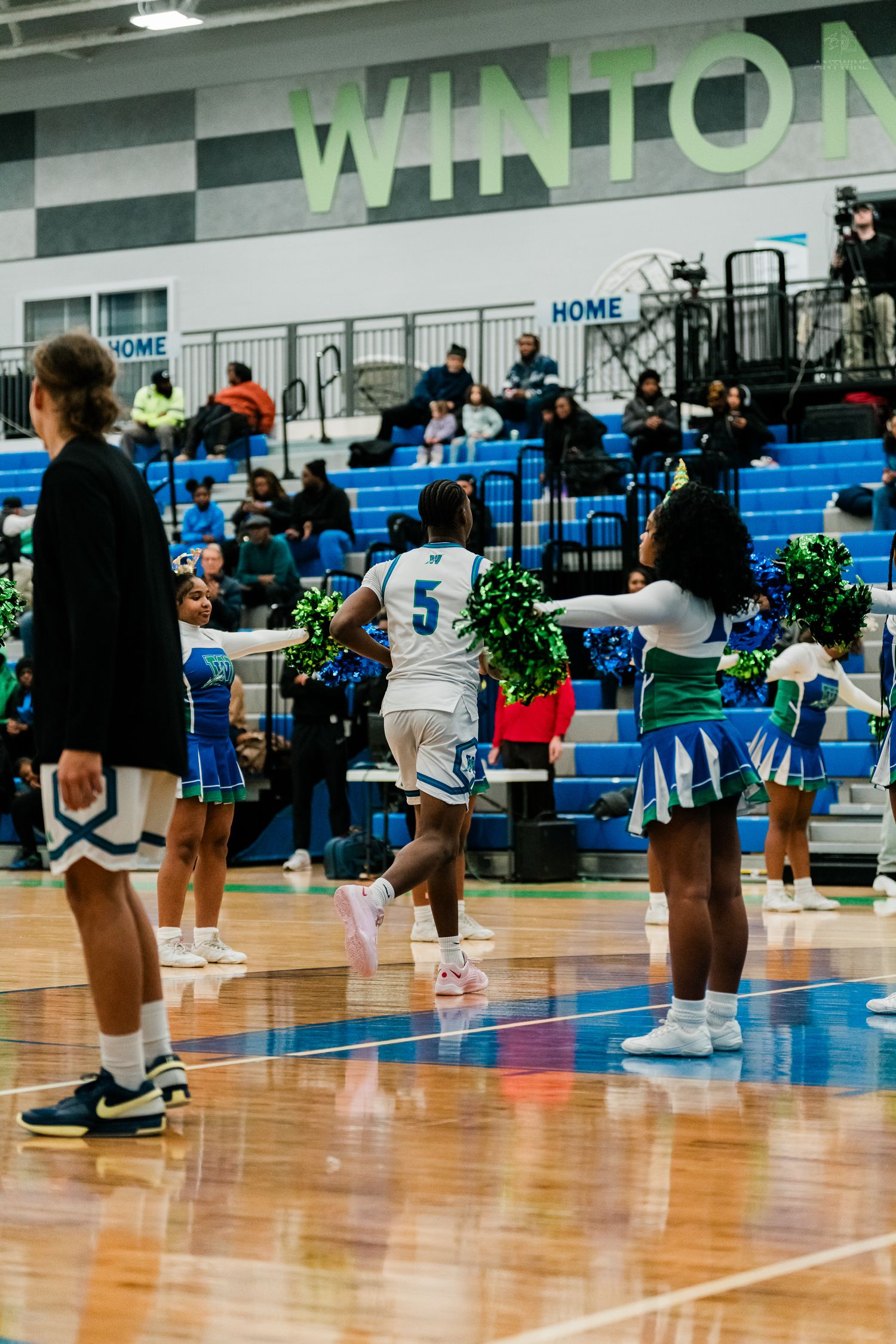 Basketball player #5 runs past cheerleaders with blue and green pom-poms at Winton school gym.