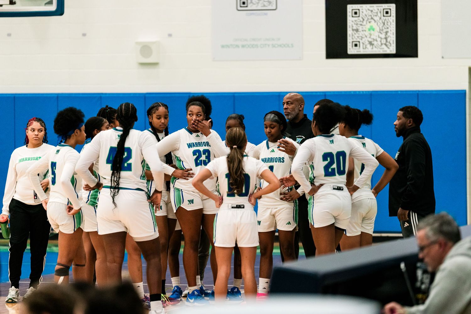 The Winton Warriors girls basketball team huddles with coaches during a timeout in a high school game.