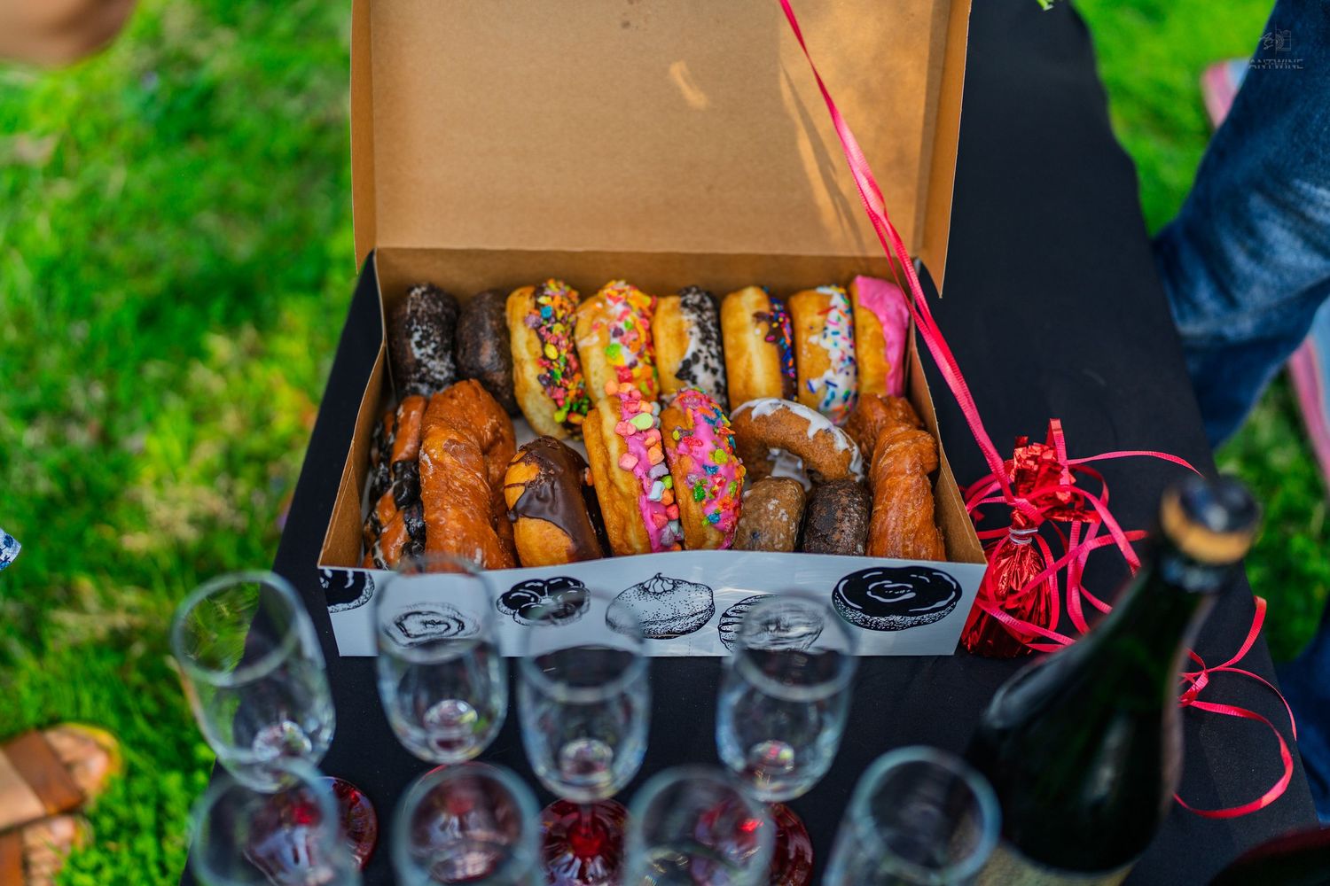 Colorful assorted decorated donuts in a black box displayed with champagne flutes on a dark table outdoors.