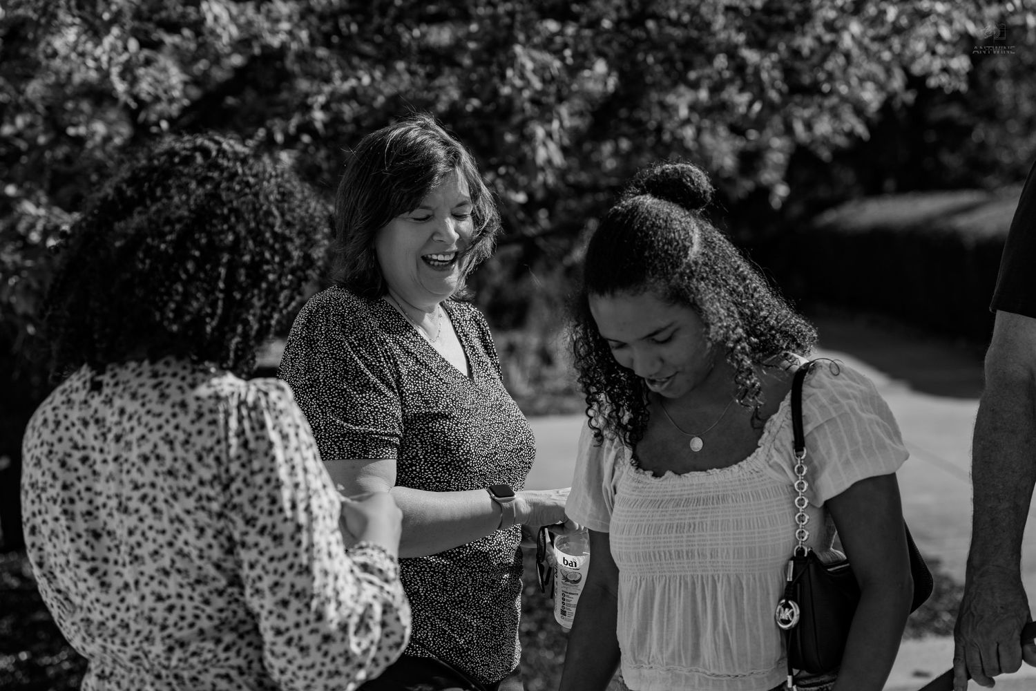 Three women laugh together outdoors in black and white photo, sharing a joyful moment in a garden setting.