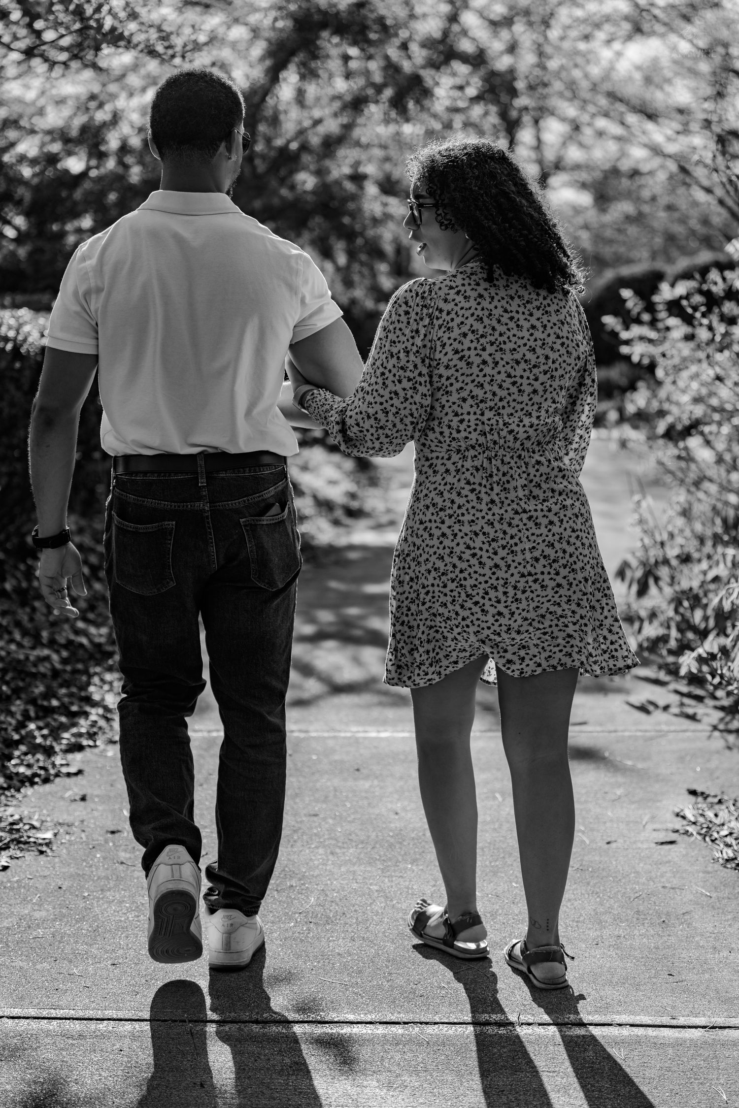 Black and white photo of a couple holding hands walking down a sunny park path.