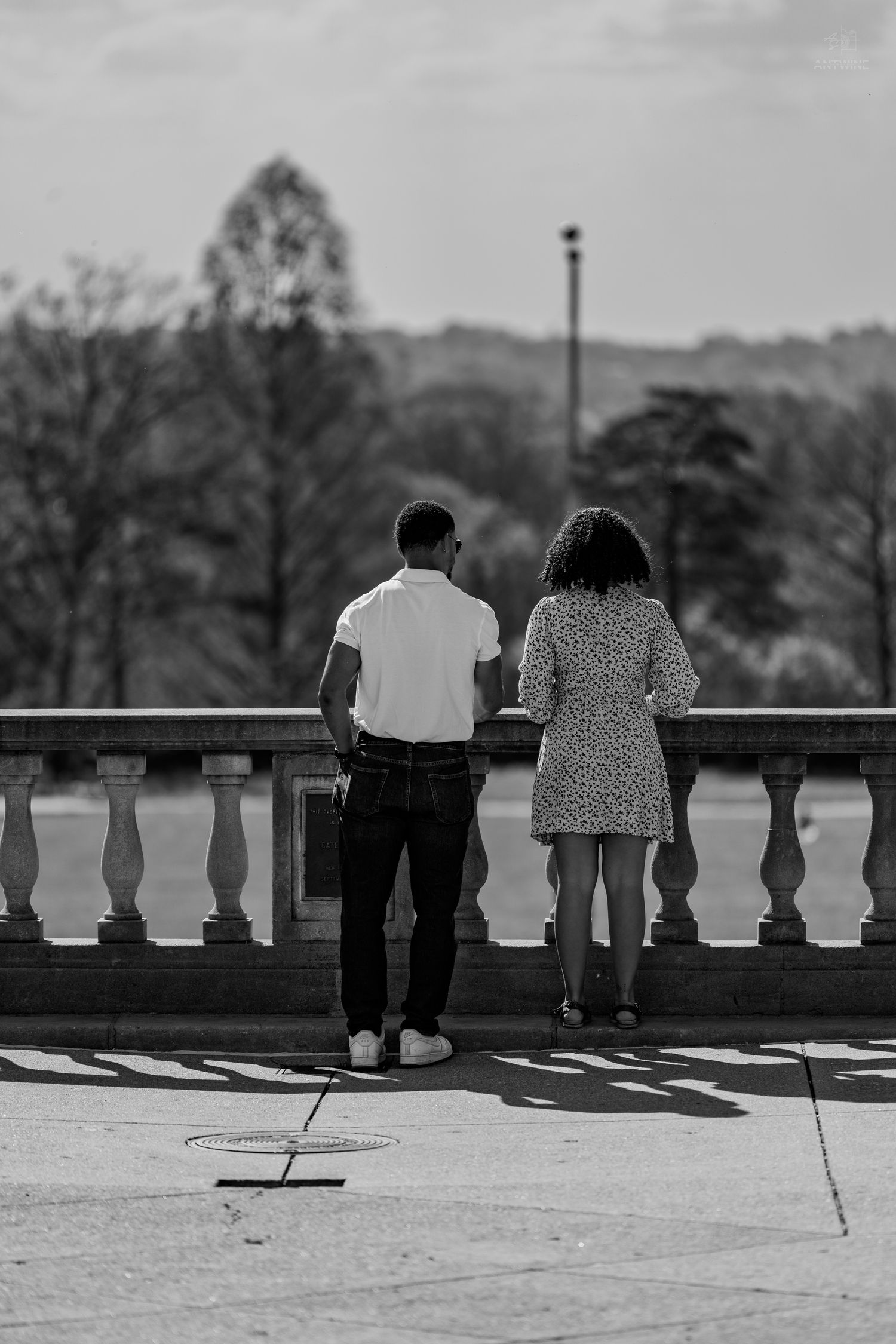 Black and white photo of couple standing at ornate stone railing overlooking park.