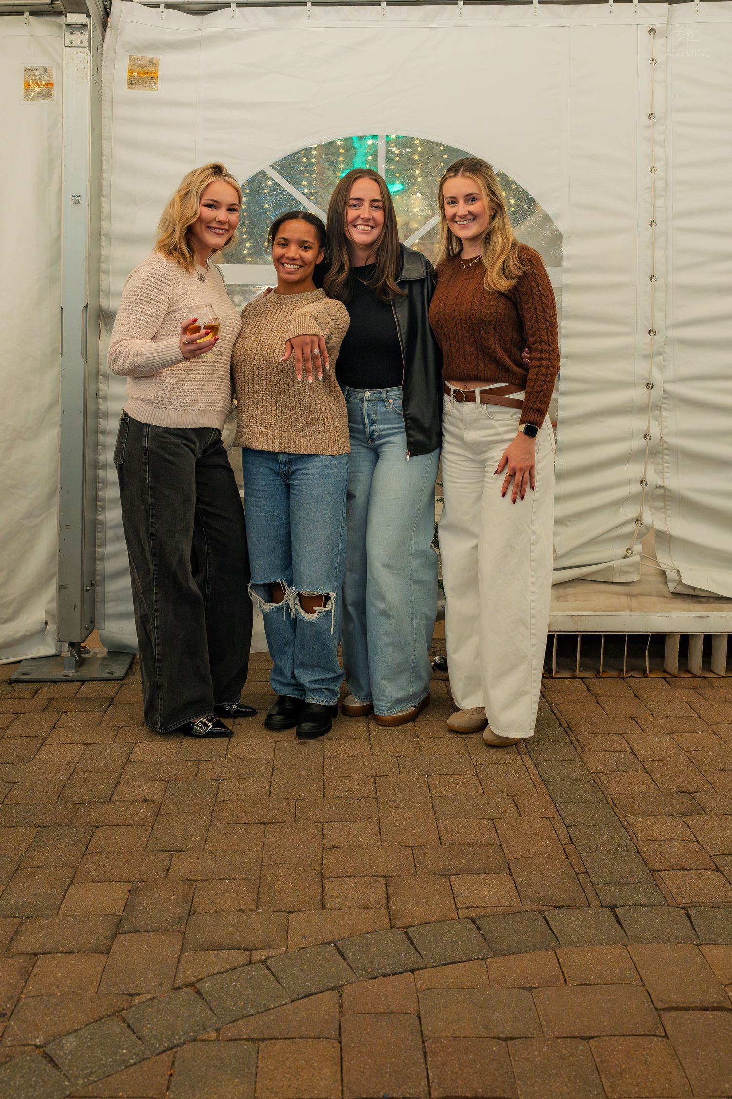 Four smiling women posing in front of a white tent wall on a brick patio at an evening event.