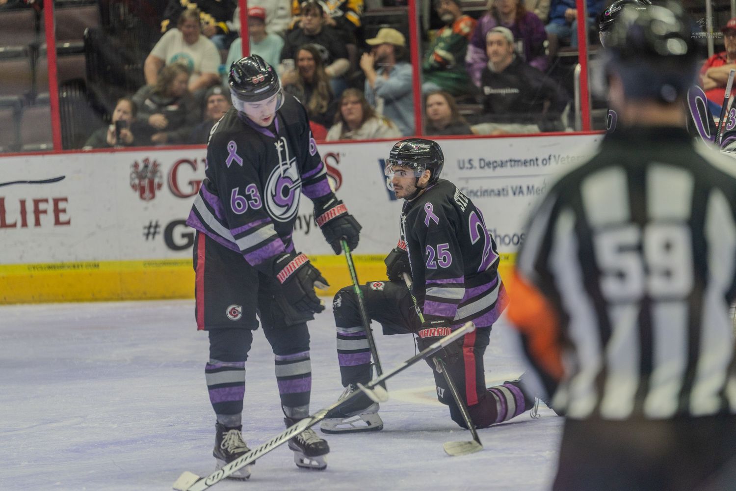 Two hockey players #63 and #25 in purple jerseys pause near referee #59 during a minor league hockey game.