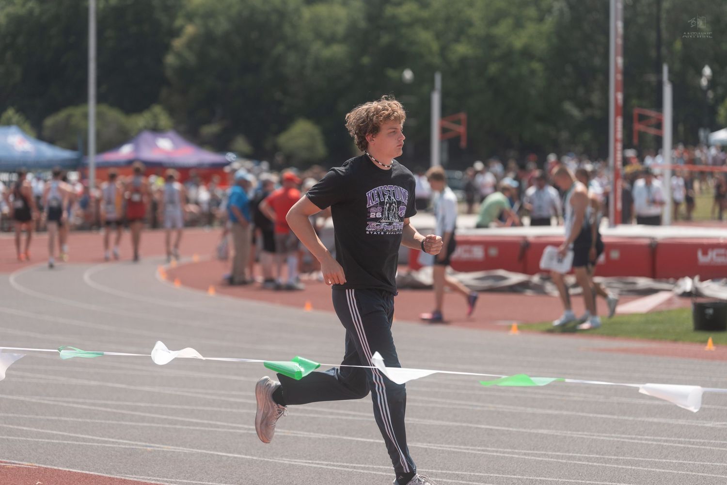 Young runner in black crosses finish line flags at sunny outdoor track and field meet.