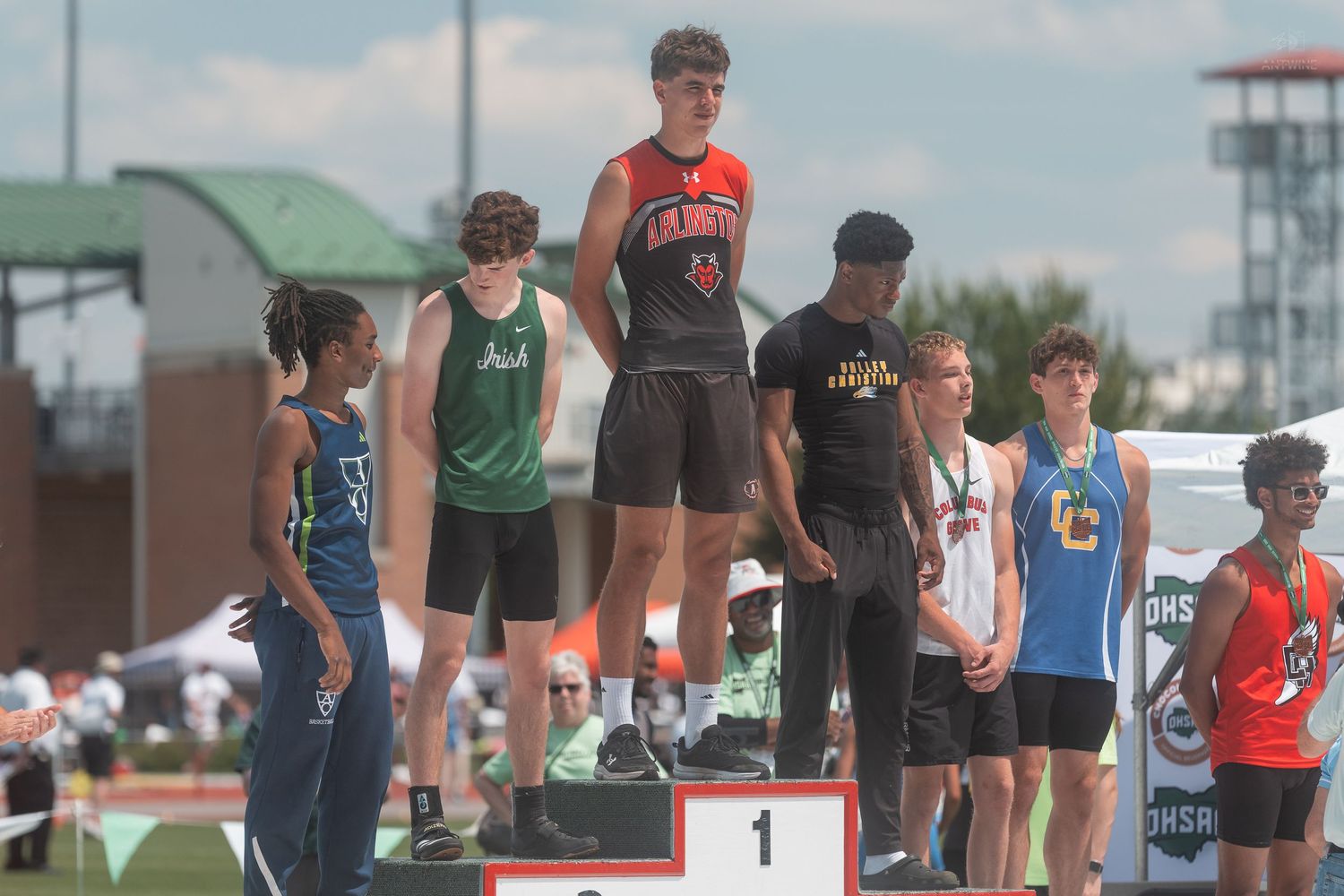 Male athlete in red jersey jumps on first place podium at OHSAA state track championship.