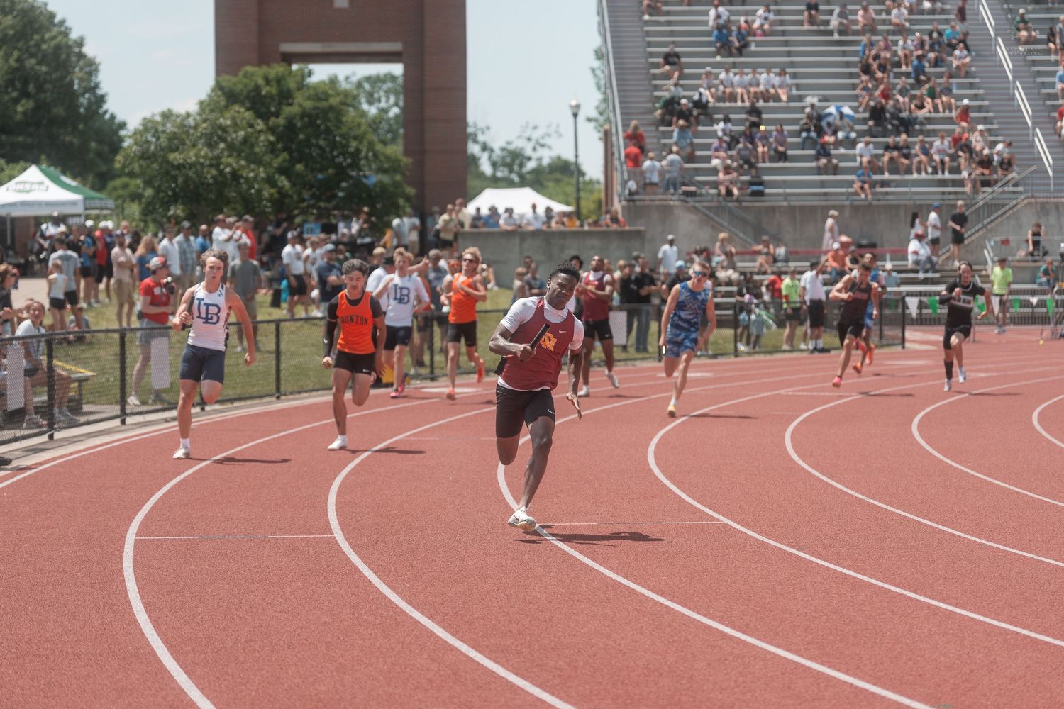 Athletes sprint on a red track during a competitive outdoor track and field meet with large crowds.