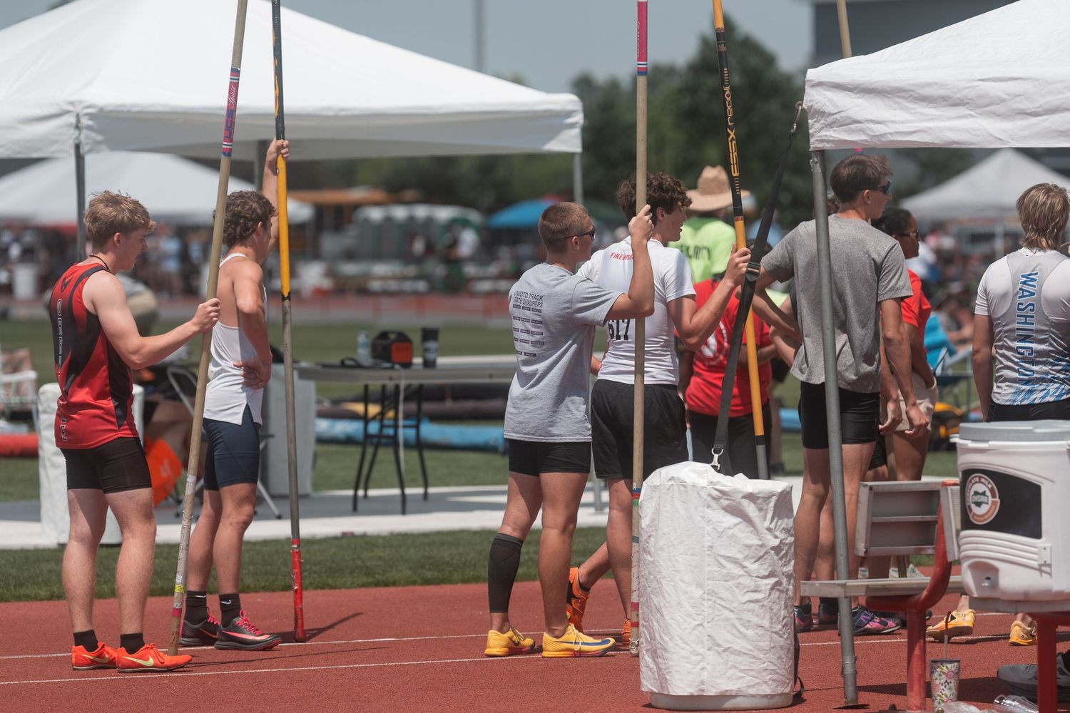 Athletes prepare for pole vault competition under white tents at an outdoor track and field meet on a sunny day.