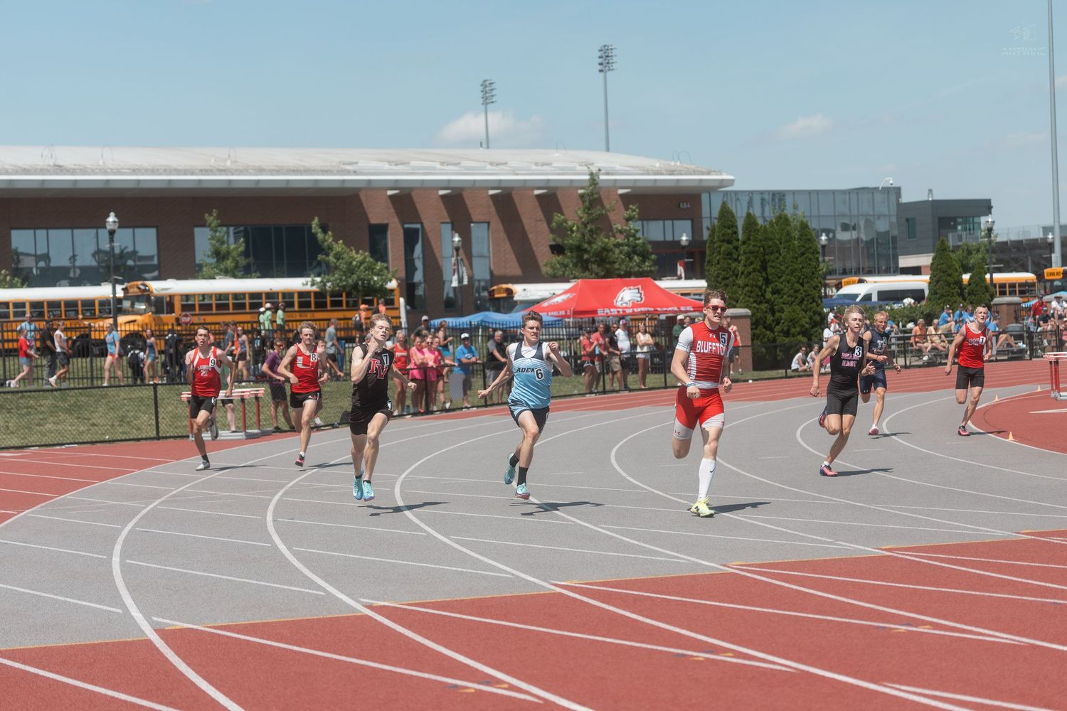 Multiple athletes racing across a red outdoor track during a high school track and field competition.
