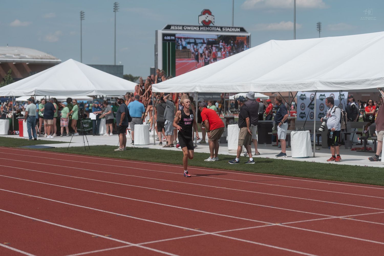 Athlete walks on red track at outdoor track and field competition with white tents and spectators in background.