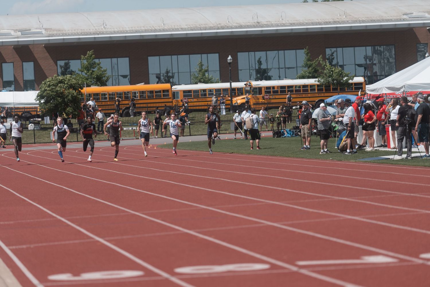 Multiple high school athletes compete in a sprint race on a red track at an outdoor track and field meet.