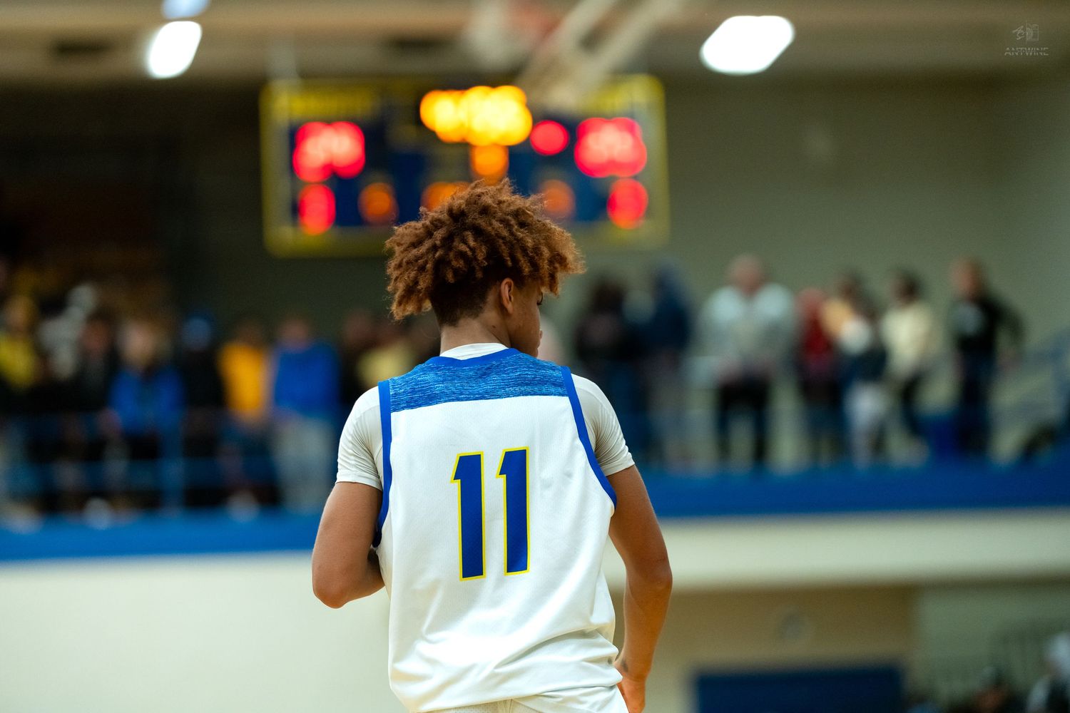 Basketball player wearing number 11 blue jersey stands near scoreboard during a game.