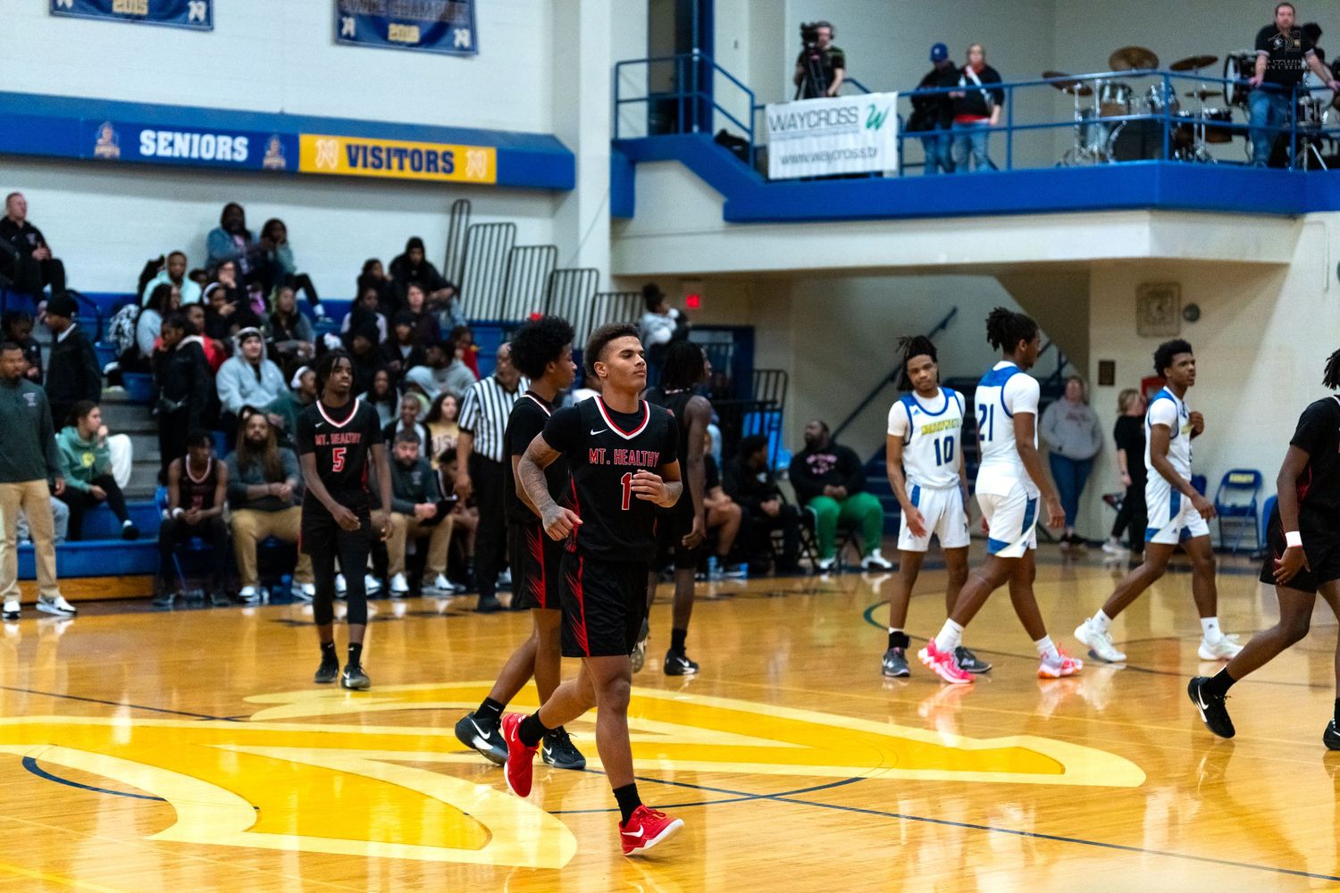 High school basketball players in red and black uniforms run down court during competitive game with packed stands.