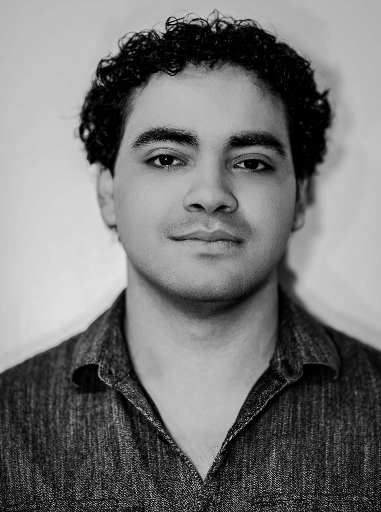 A young man with curly dark hair wearing a gray shirt poses in a black and white close-up portrait.