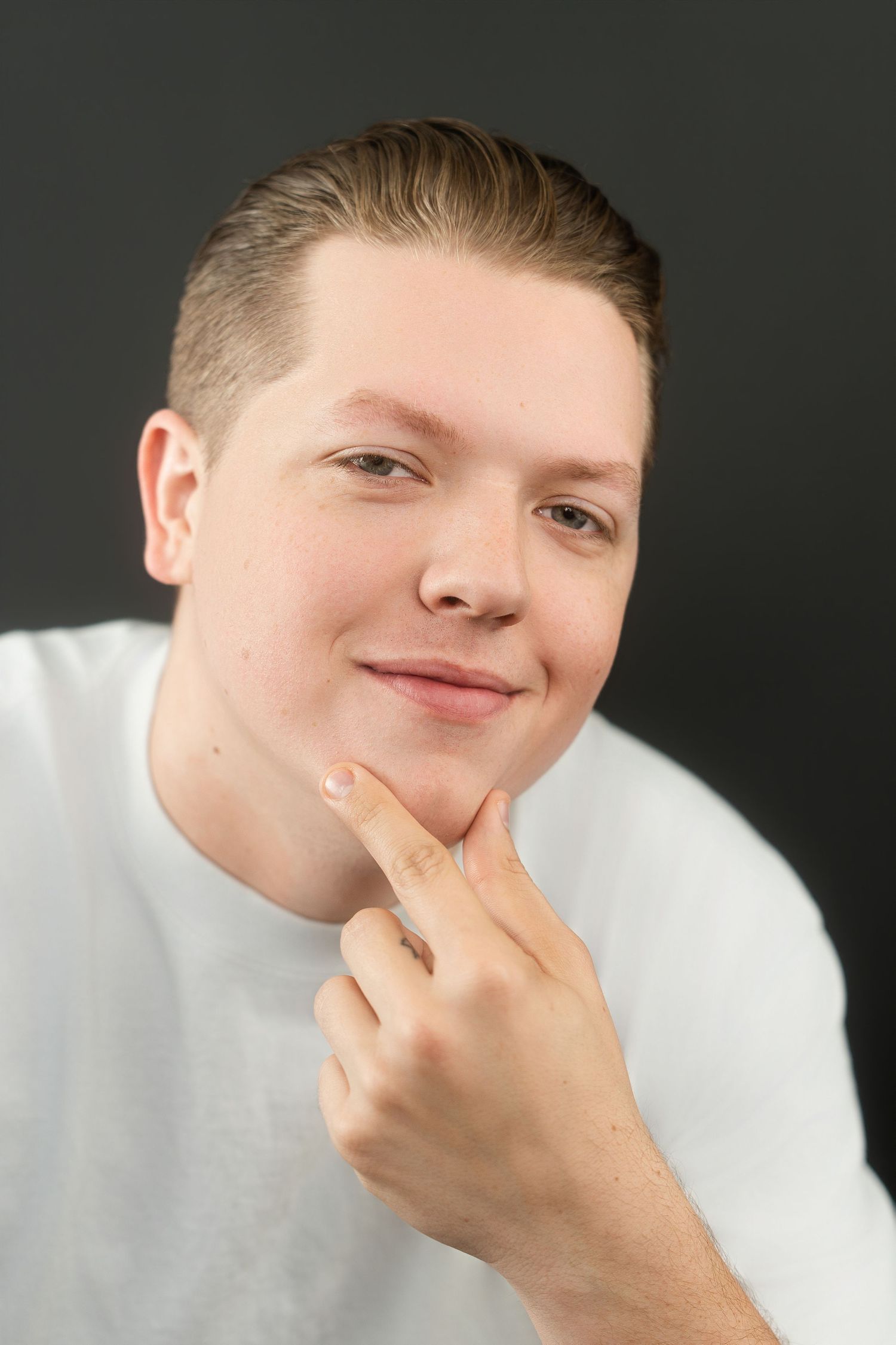 Young man in white t-shirt smiling with finger on chin in studio portrait against dark background.