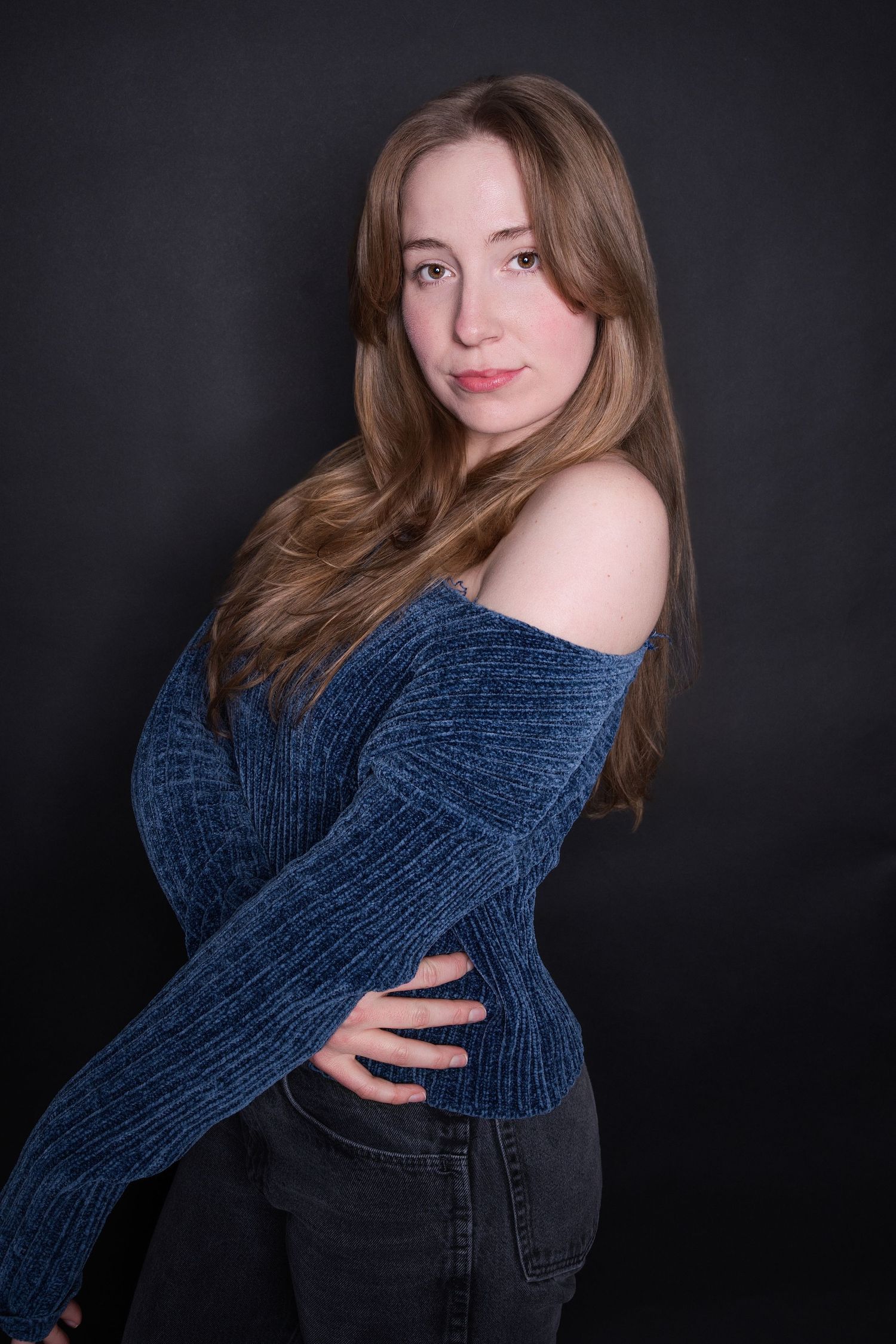 Young woman in blue off-shoulder knit sweater and jeans posing against dark background.