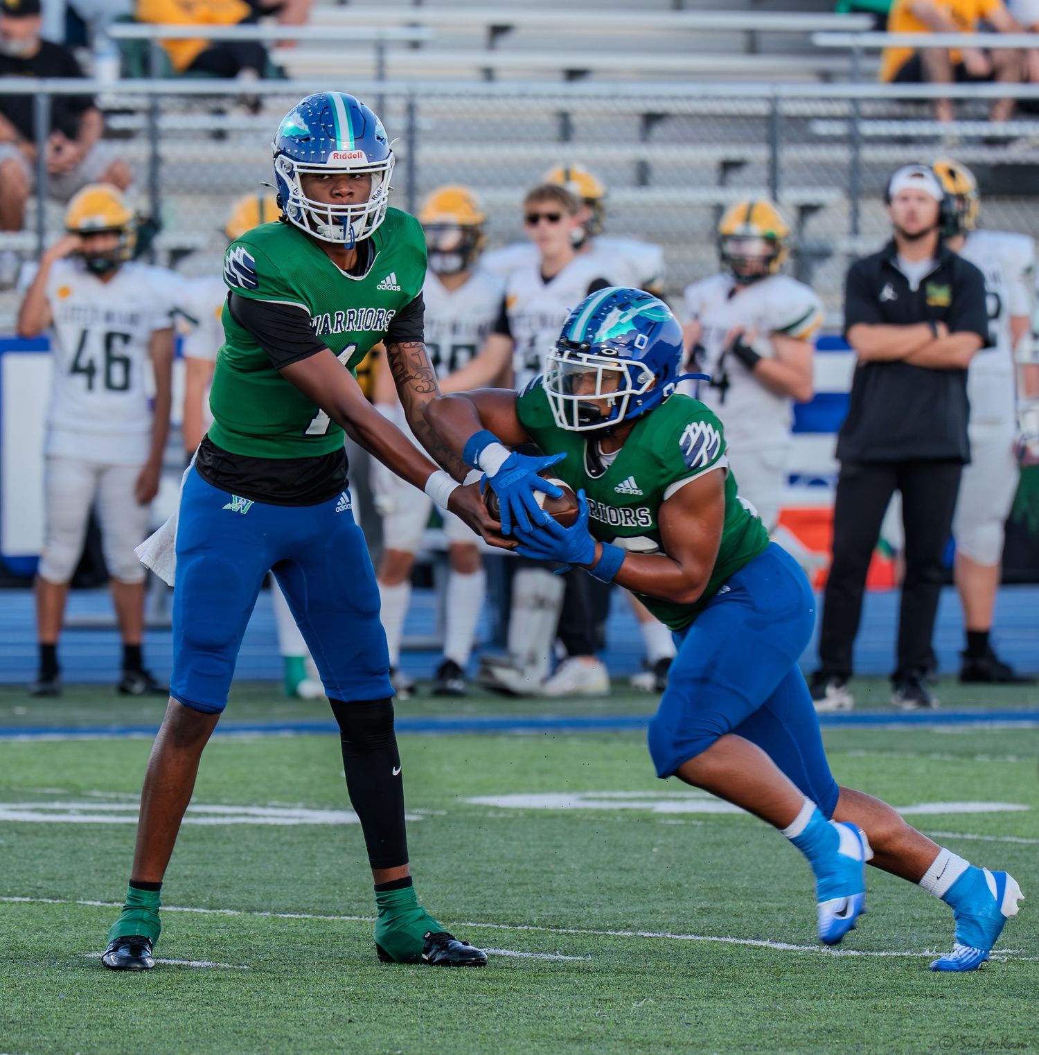 High school football quarterback hands off the ball to a running back in green and blue uniforms.