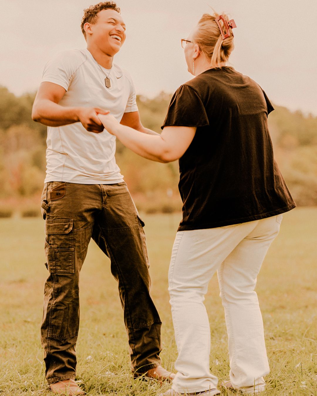 Couple laughing and dancing together in a sunny outdoor field during casual wedding celebration.