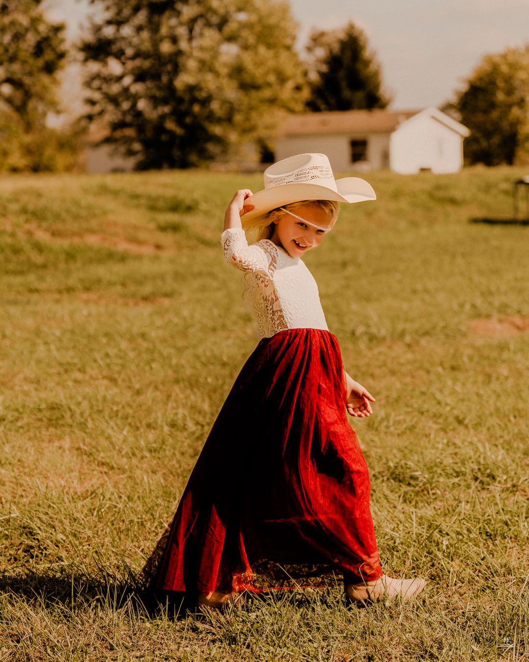 Young flower girl in red maxi skirt and white cowboy hat twirling in a sunlit green field.