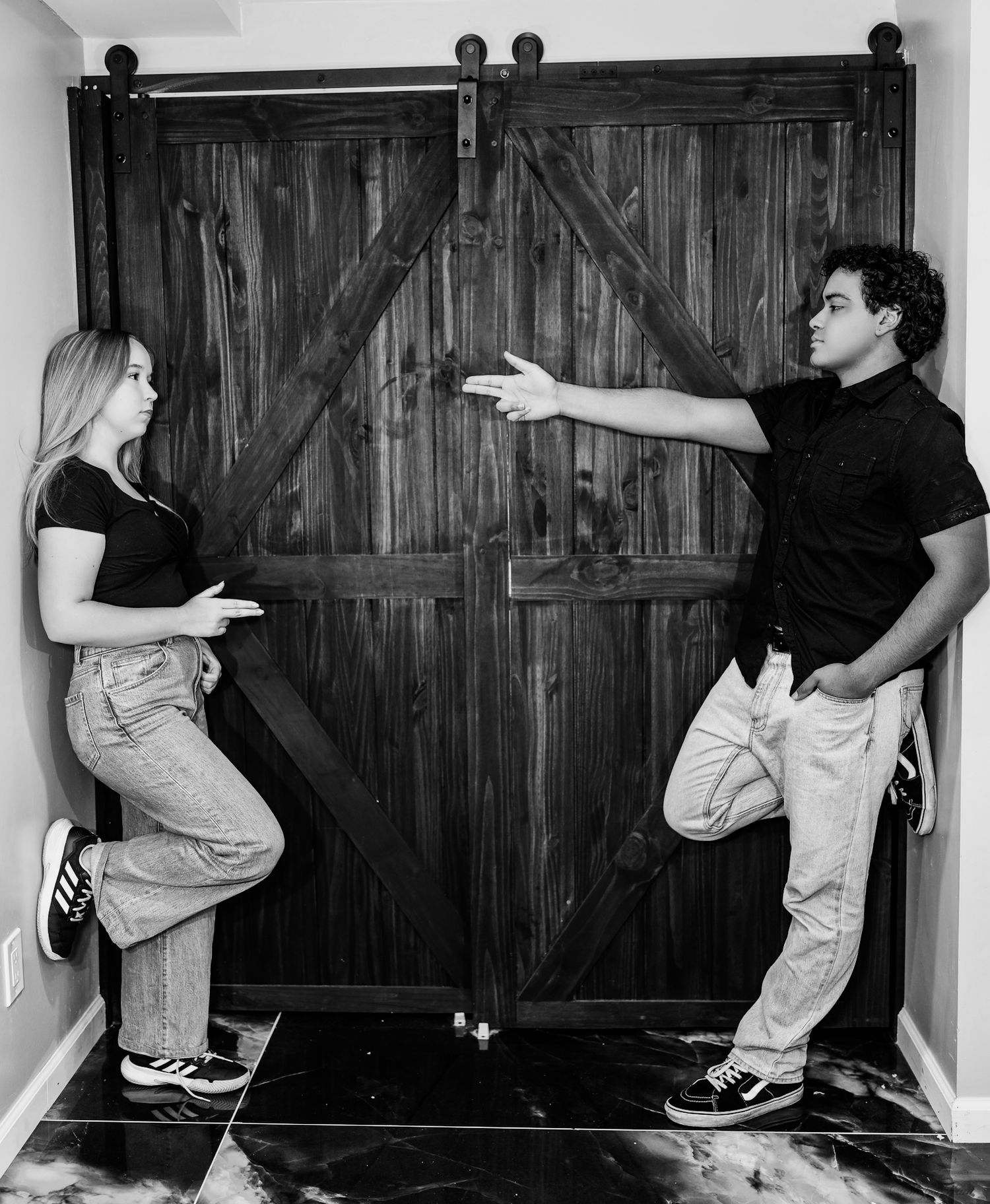 Black and white photo of young couple facing each other by rustic barn door, man gesturing.