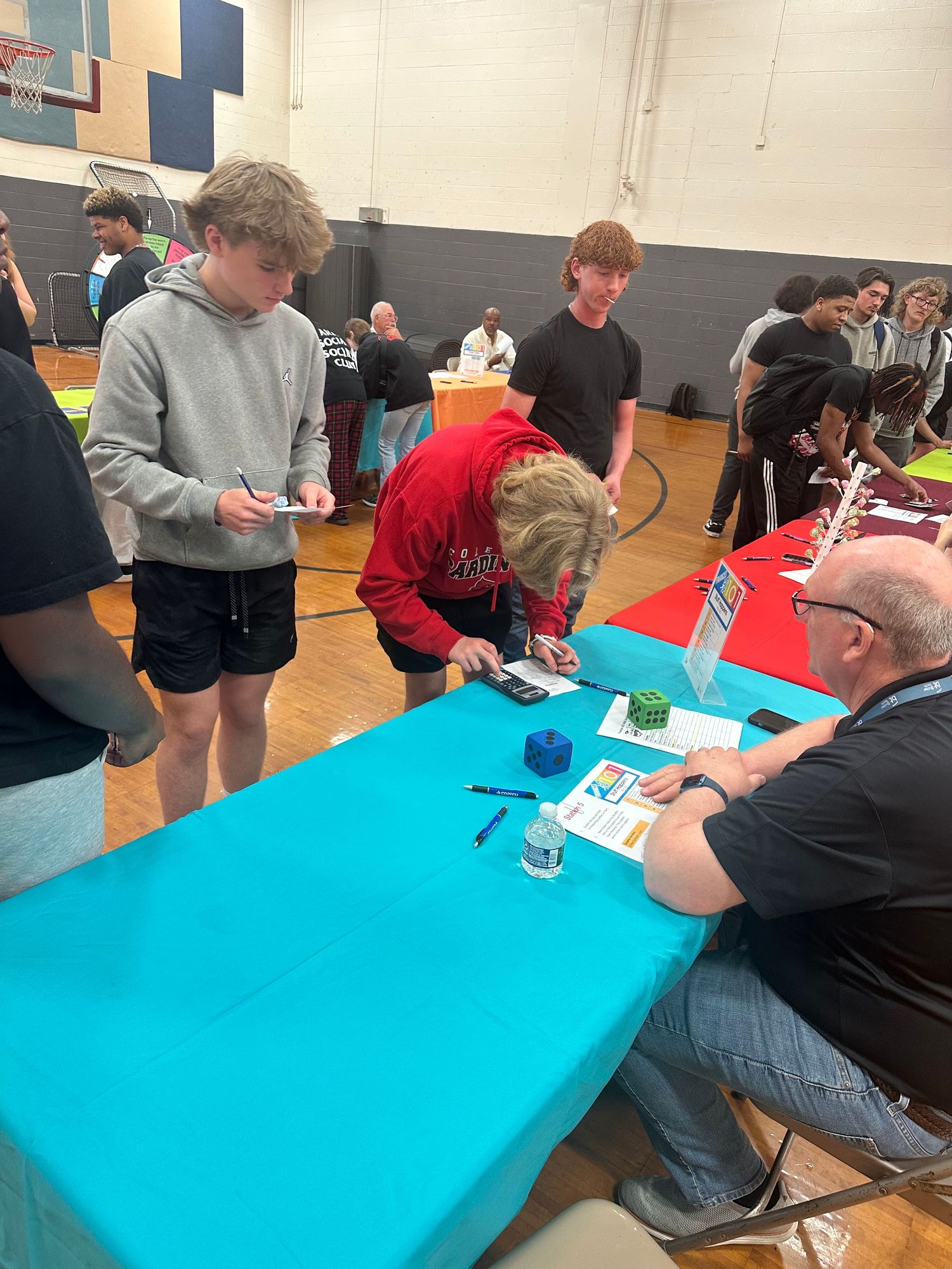 Students sign up at a teal table with a seated representative during a school gymnasium career fair.