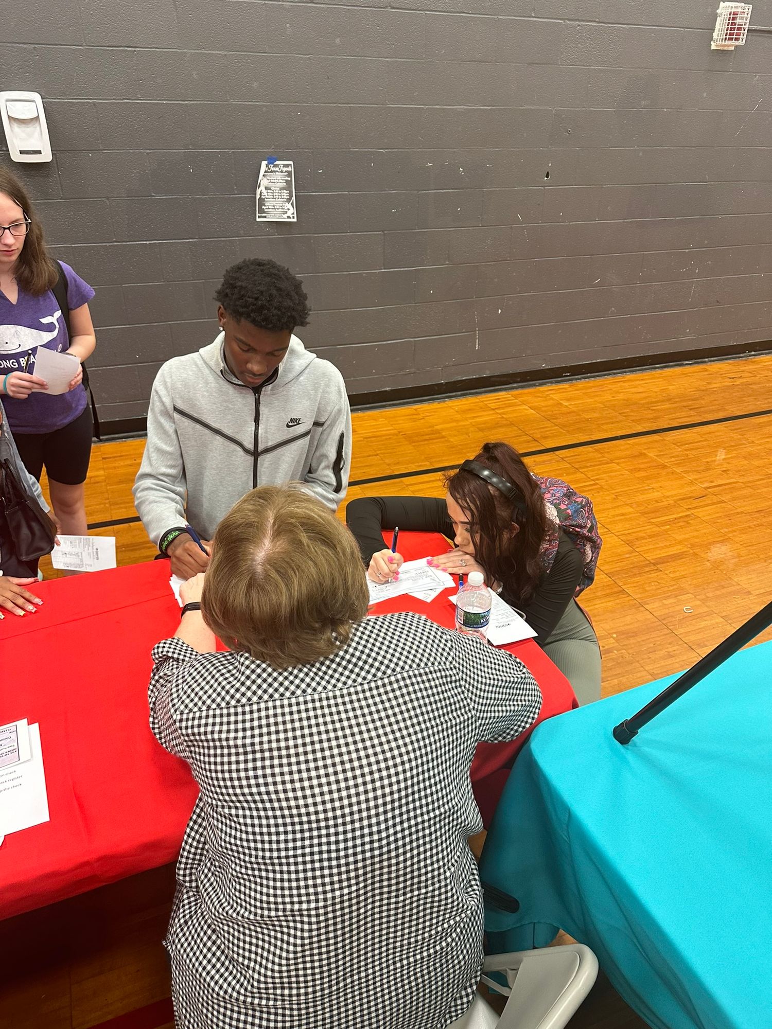Students and adults collaborate over paperwork at a red table during an indoor school fair event.