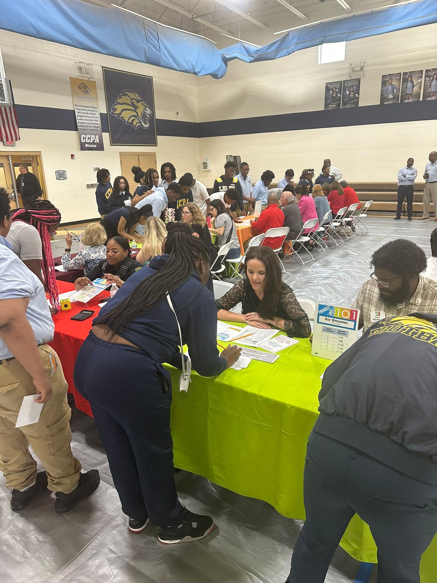 People gathered at tables in a school gymnasium for a community job fair event with bright green and red tablecloths.