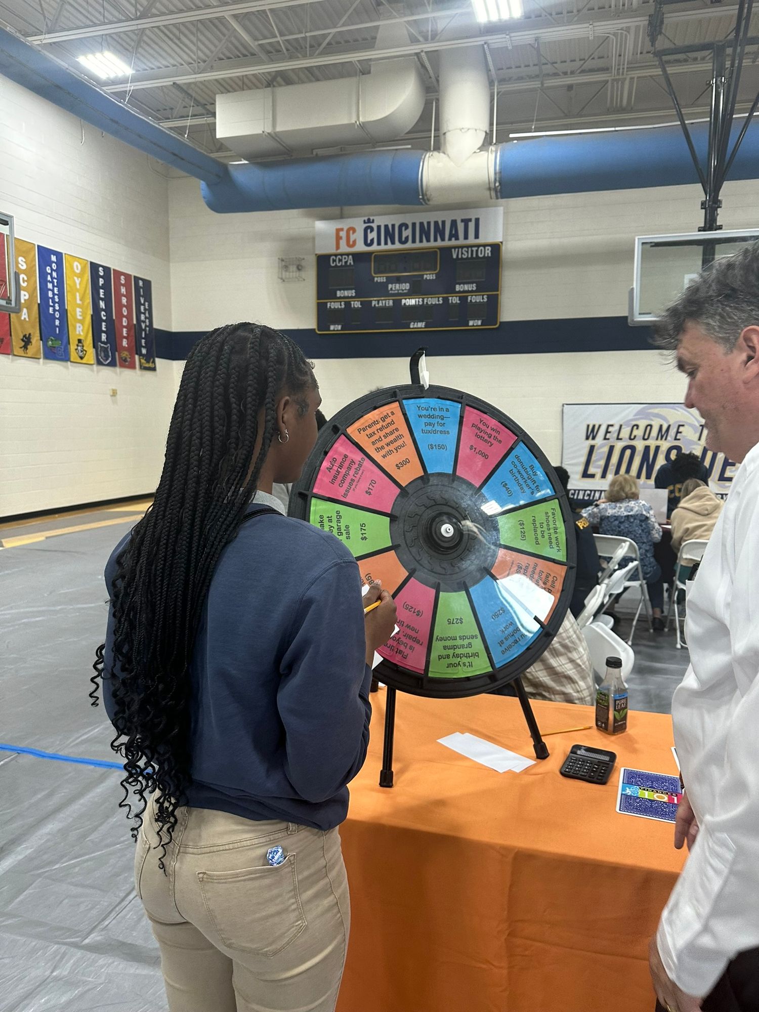 A student spins a colorful prize wheel at a school gymnasium event with FC Cincinnati signage visible.