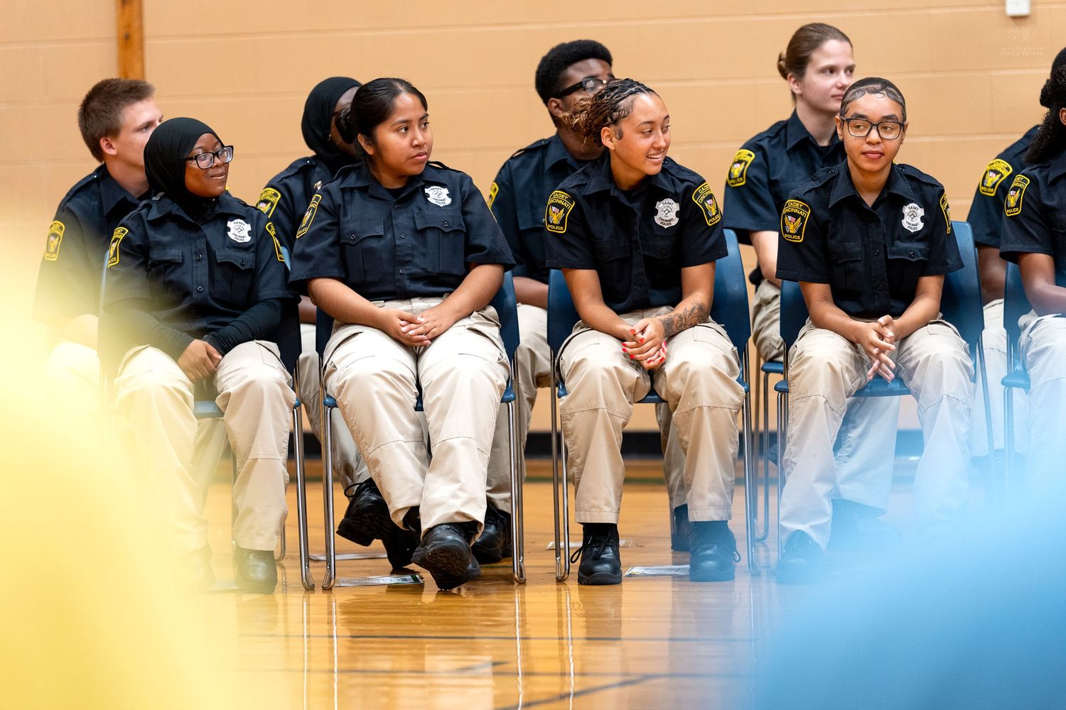 Young cadets in uniform shirts and khaki pants sit together in a gymnasium during a formal ceremony or assembly.