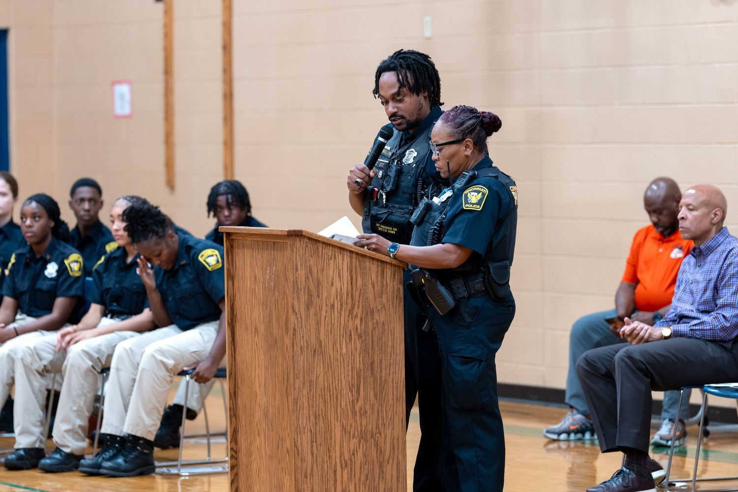Two police officers speak at a podium during a community meeting in a gymnasium with attendees seated in the background.
