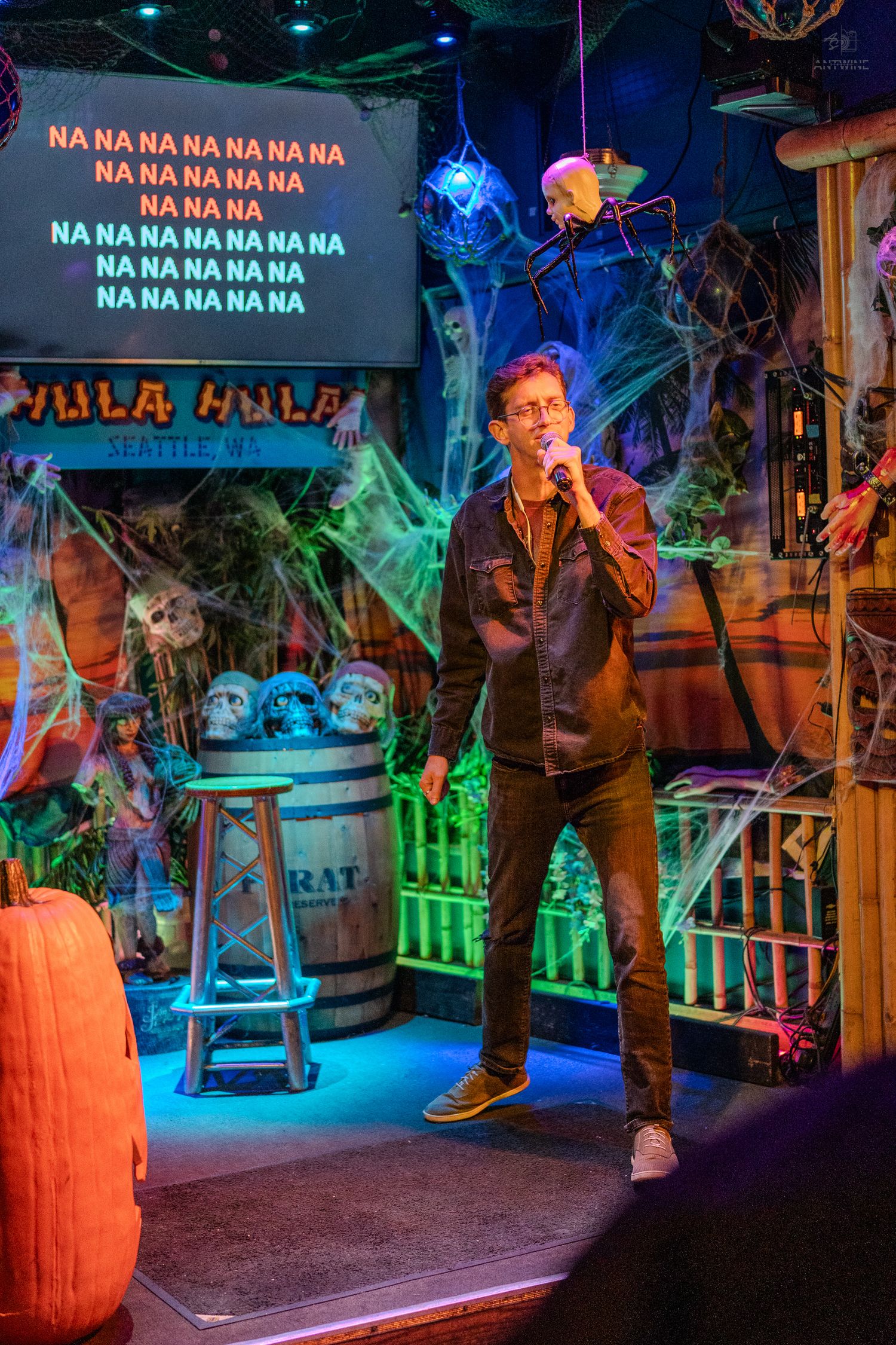 Man singing karaoke on Halloween-decorated stage at Hula Hula bar in Seattle, with spooky cobwebs and skulls.