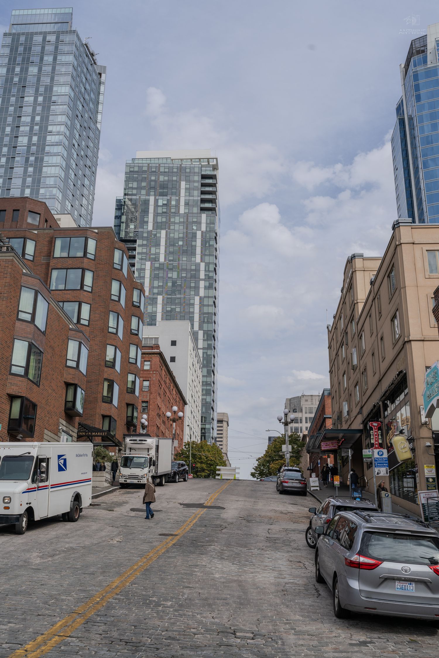 Urban street scene with USPS truck, modern skyscrapers, brick buildings, and parked cars on a cobblestone road under cloudy skies.