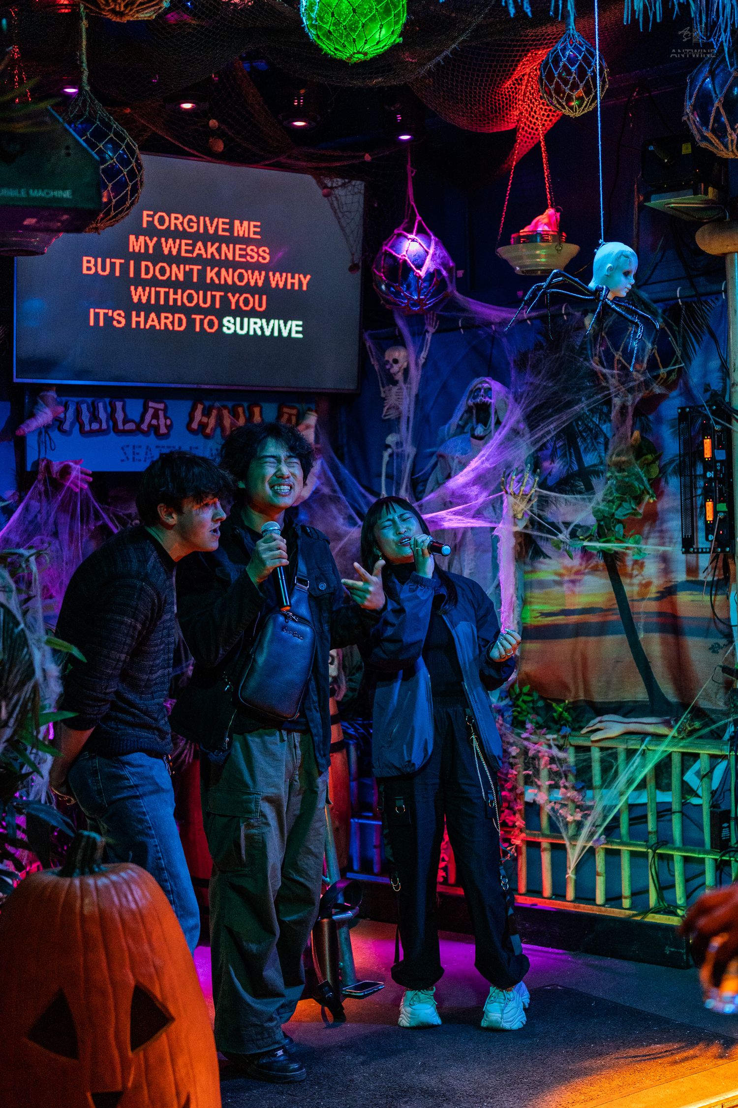 Three friends singing karaoke on a Halloween-decorated stage with colorful neon lighting.