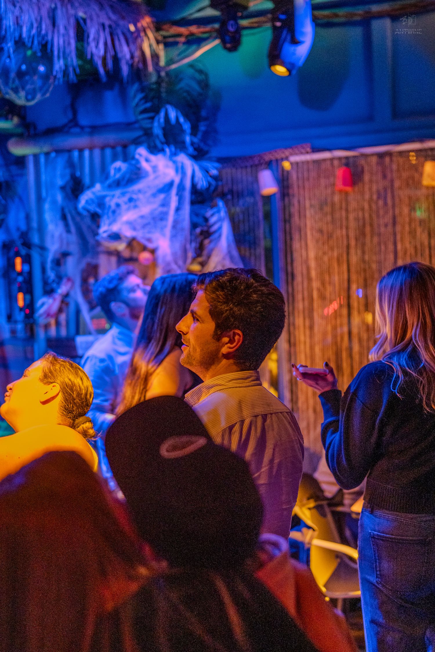 People socializing in a dimly lit tropical-themed bar with blue and orange neon lighting.