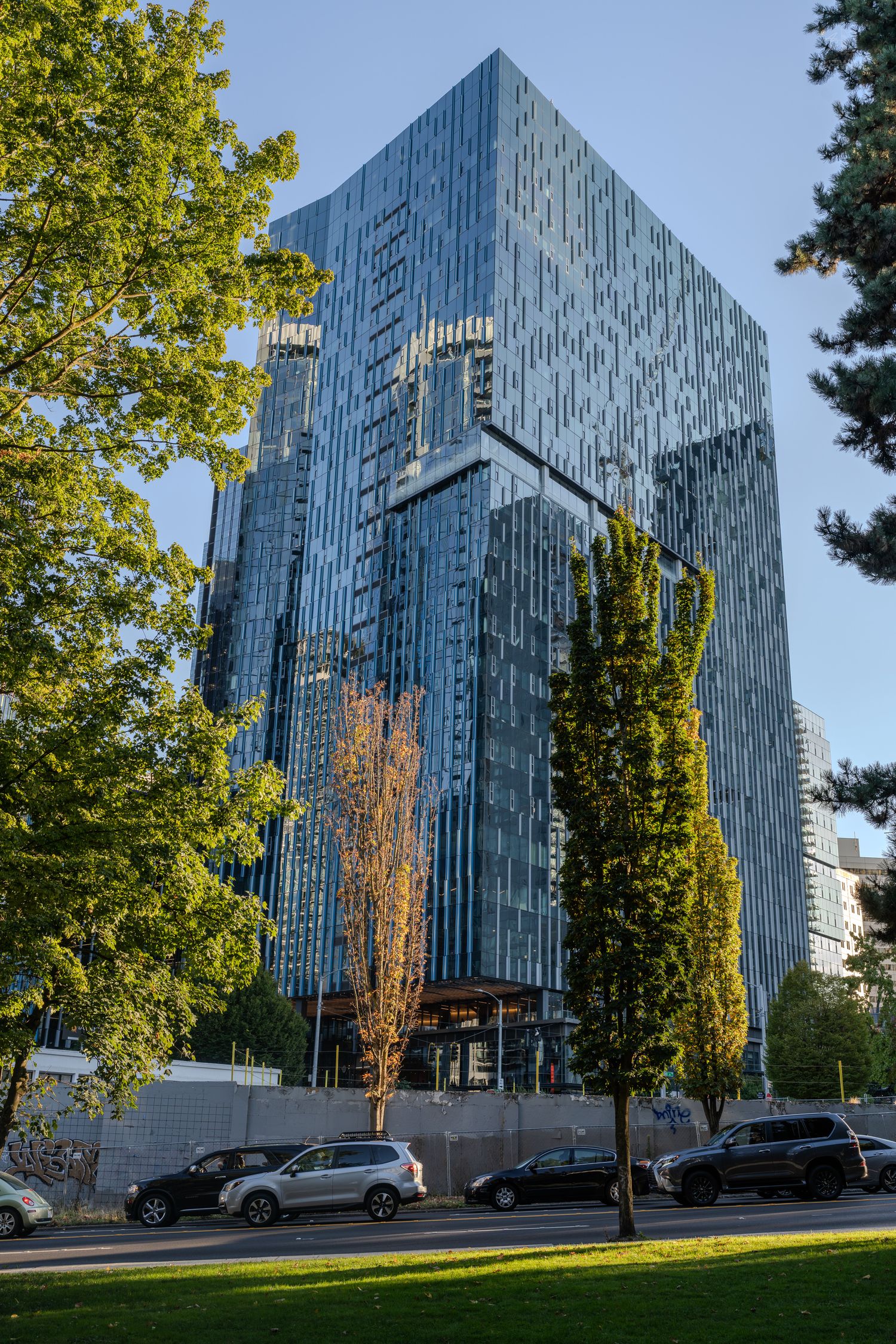 Modern glass skyscraper reflecting blue sky surrounded by green trees in urban setting.