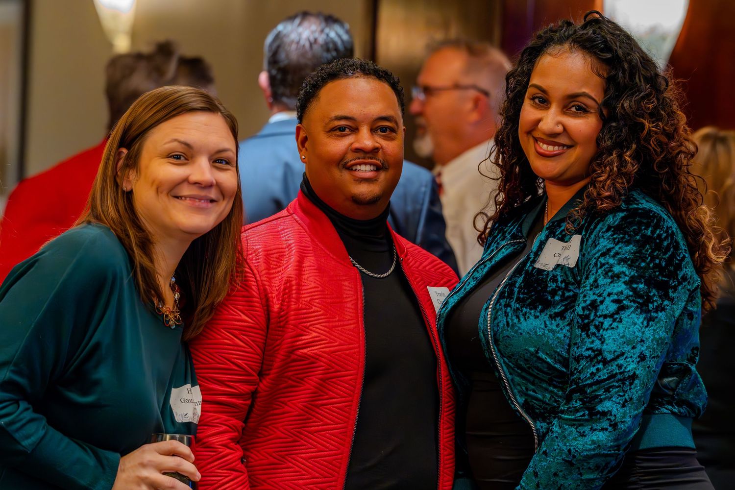 Three smiling people pose together at a social event, wearing colorful attire including red and teal jackets.
