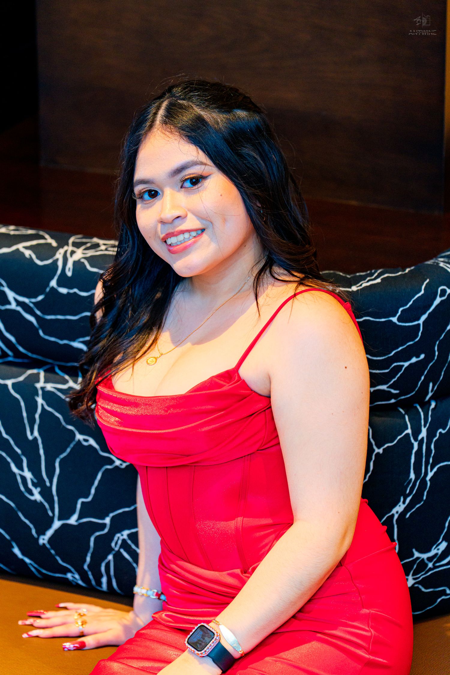 Smiling young woman in red spaghetti strap dress seated on dark sofa wearing a smartwatch.