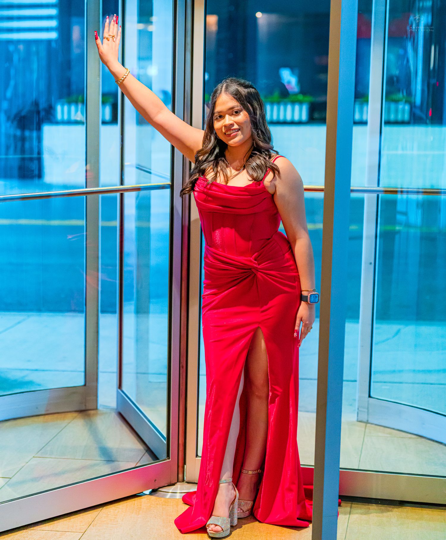 Woman in red velvet slit gown waves while posing at glass entrance doors.