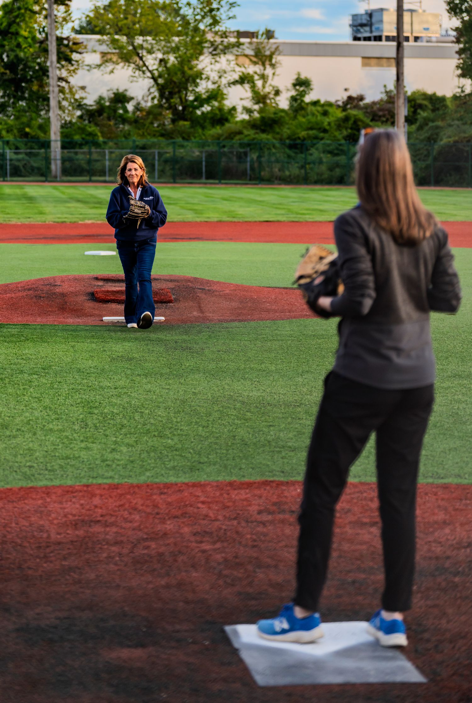Woman stands on pitcher's mound holding softball while another player waits at home plate on green turf field.