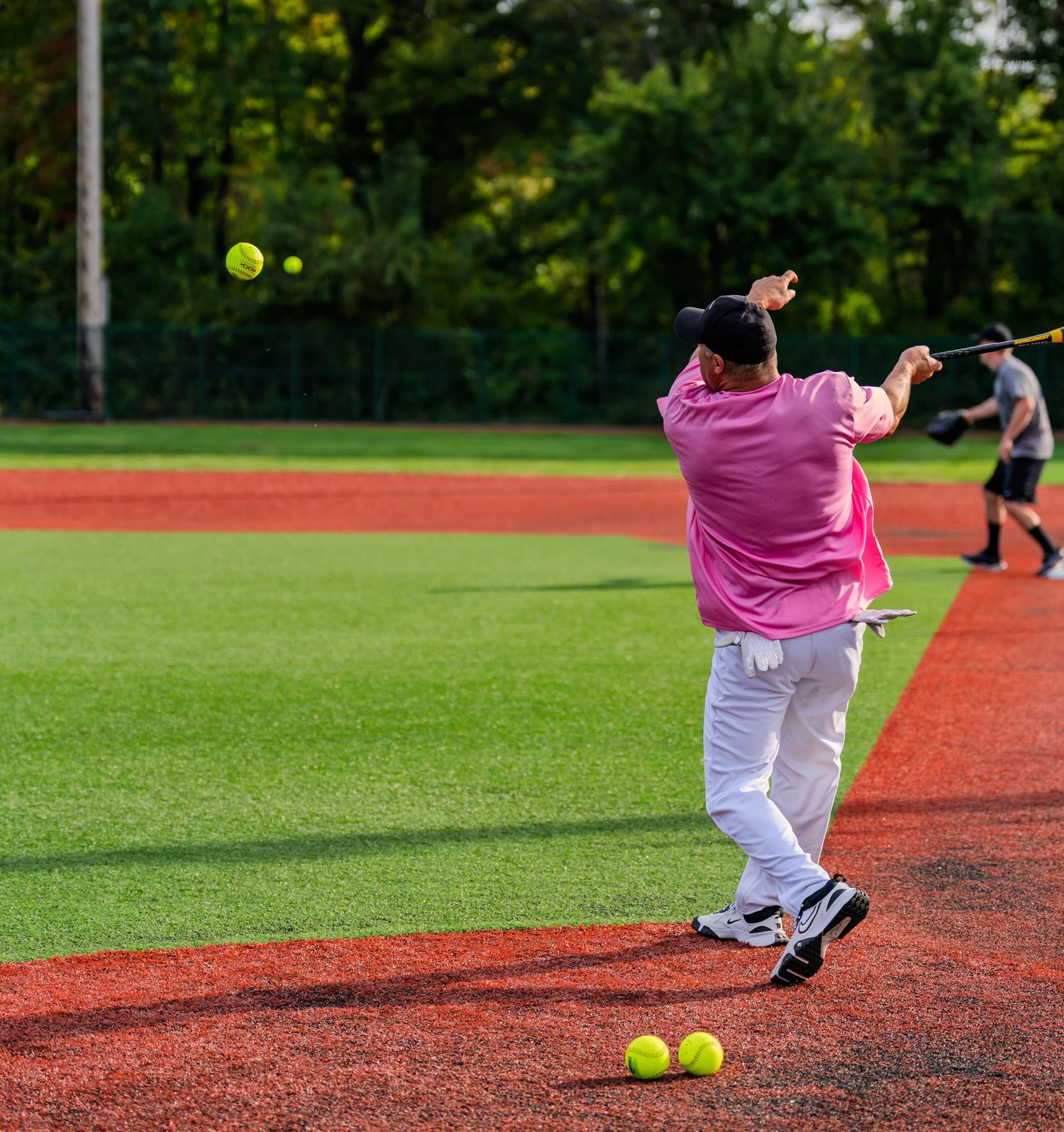 Person in pink shirt hitting softballs on a baseball field during batting practice with yellow balls on ground.