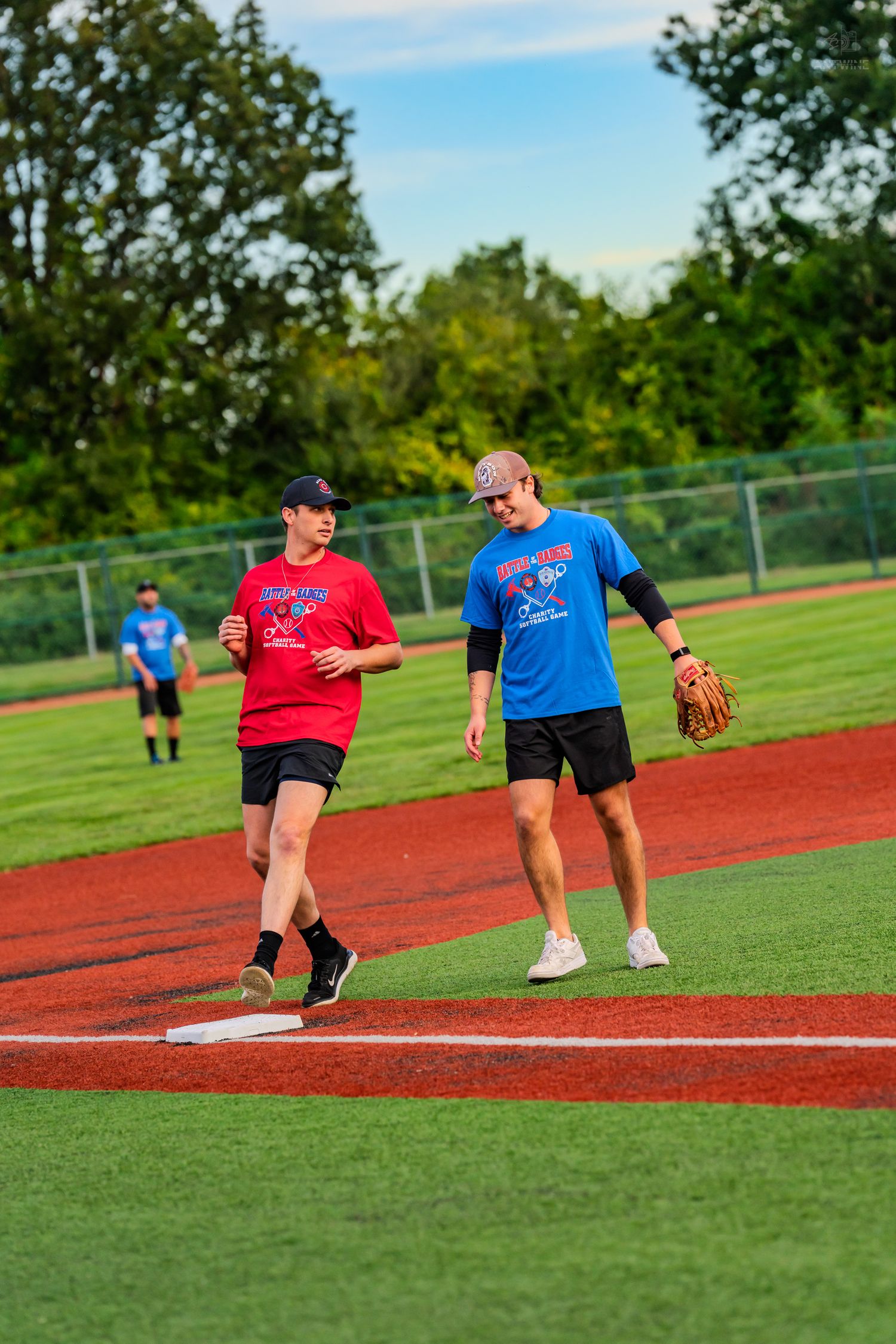 Two players in red and blue shirts chat near a base on a green softball field.