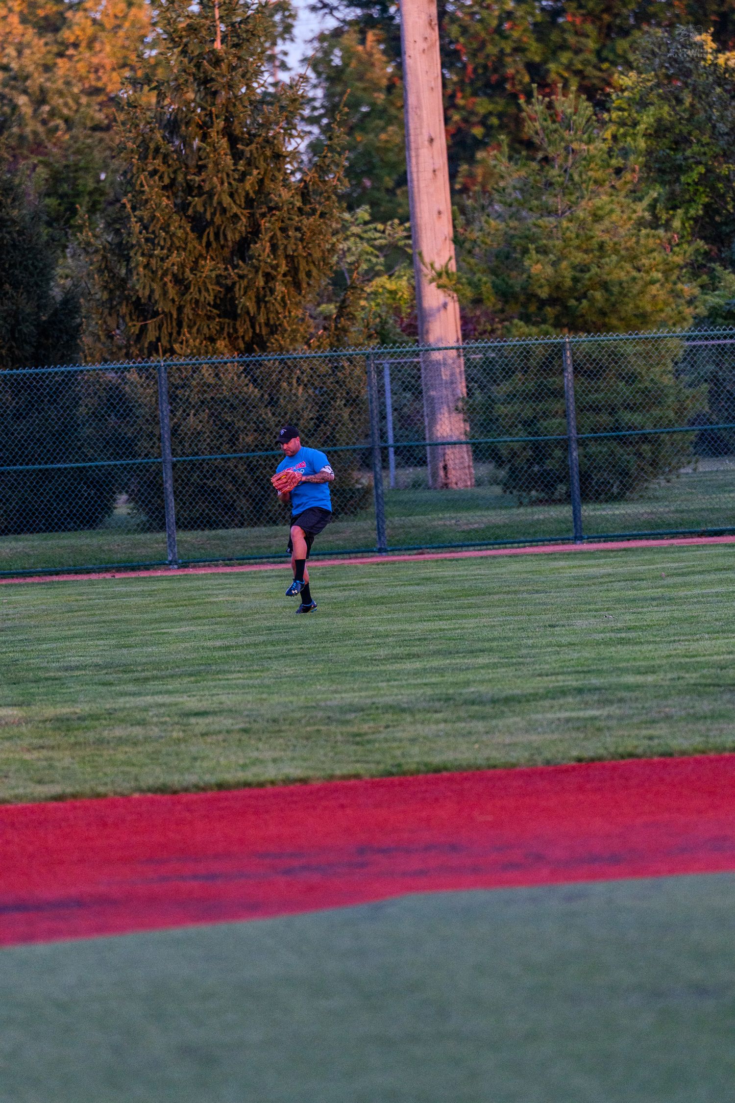Outfielder in blue jersey runs to field a ball during an evening softball game on a well-maintained diamond.