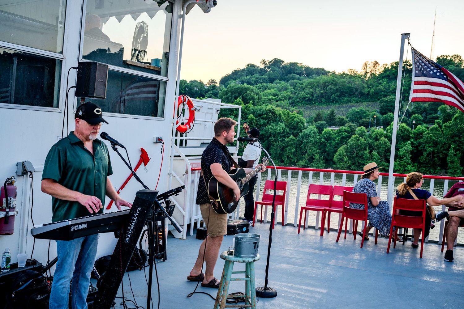 Live band performing on a riverboat deck at sunset with an American flag and scenic green hills in the background.