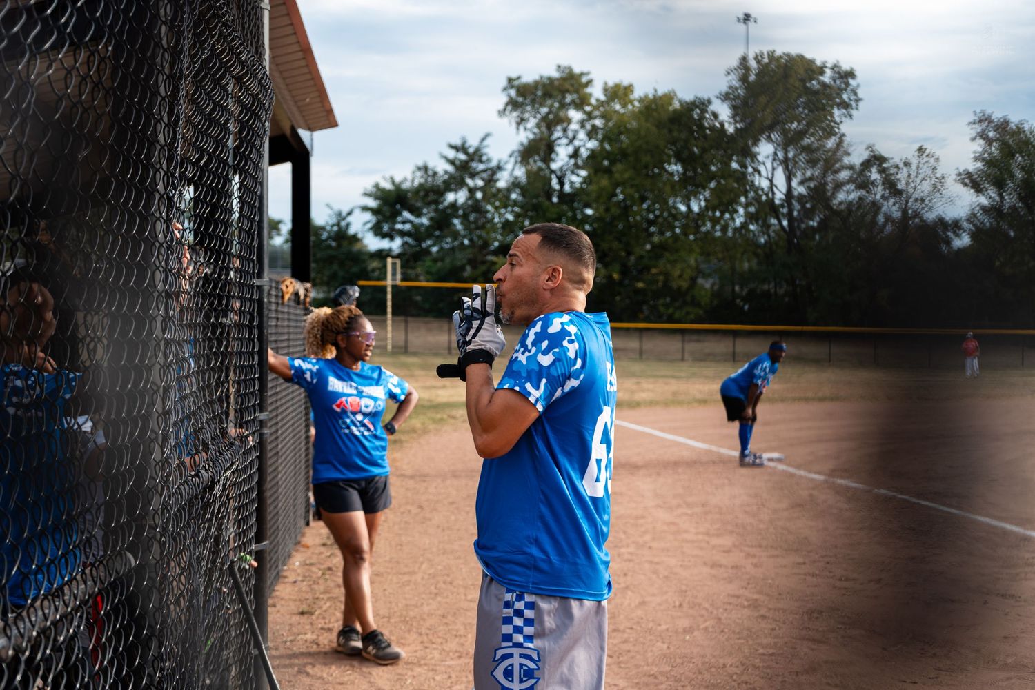 A youth baseball player in a blue jersey stands near a chain-link fence during a softball game outdoors.